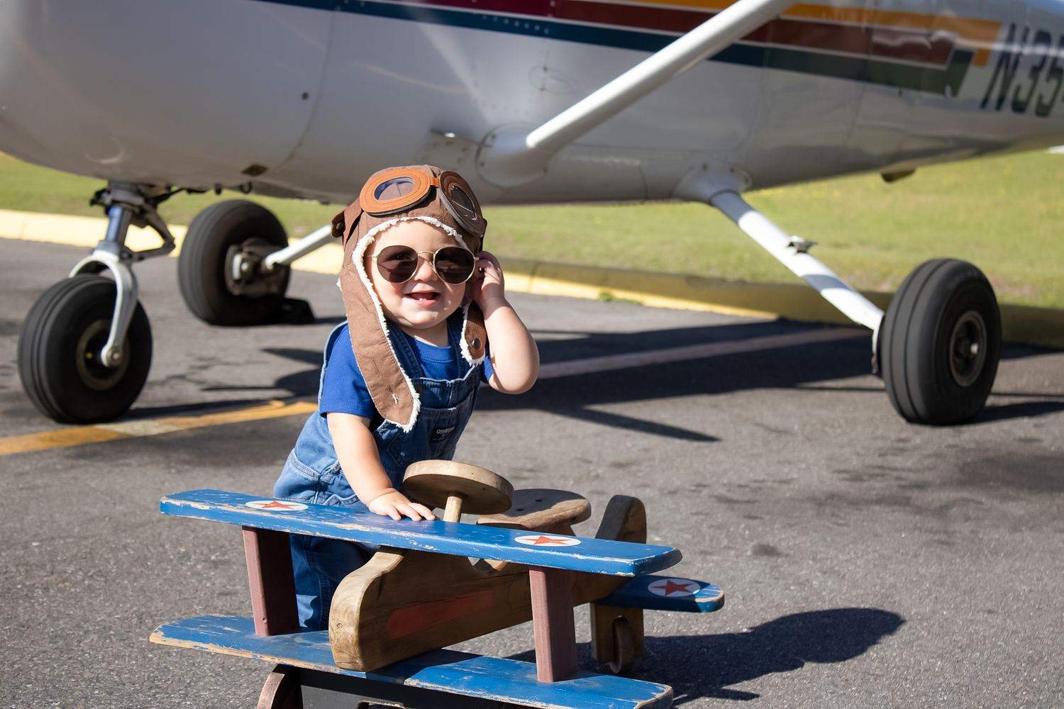 Young child dressed as a pilot with goggles and helmet sits on a toy airplane in front of a real aircraft.