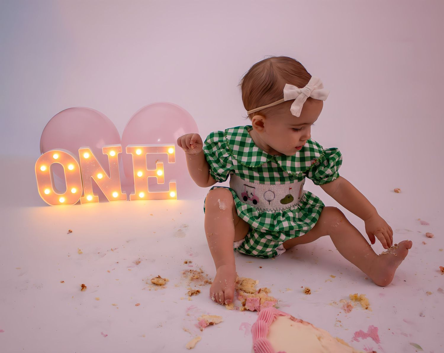 Baby girl in green gingham dress celebrates first birthday with cake smash near lit ONE sign and pink balloons.