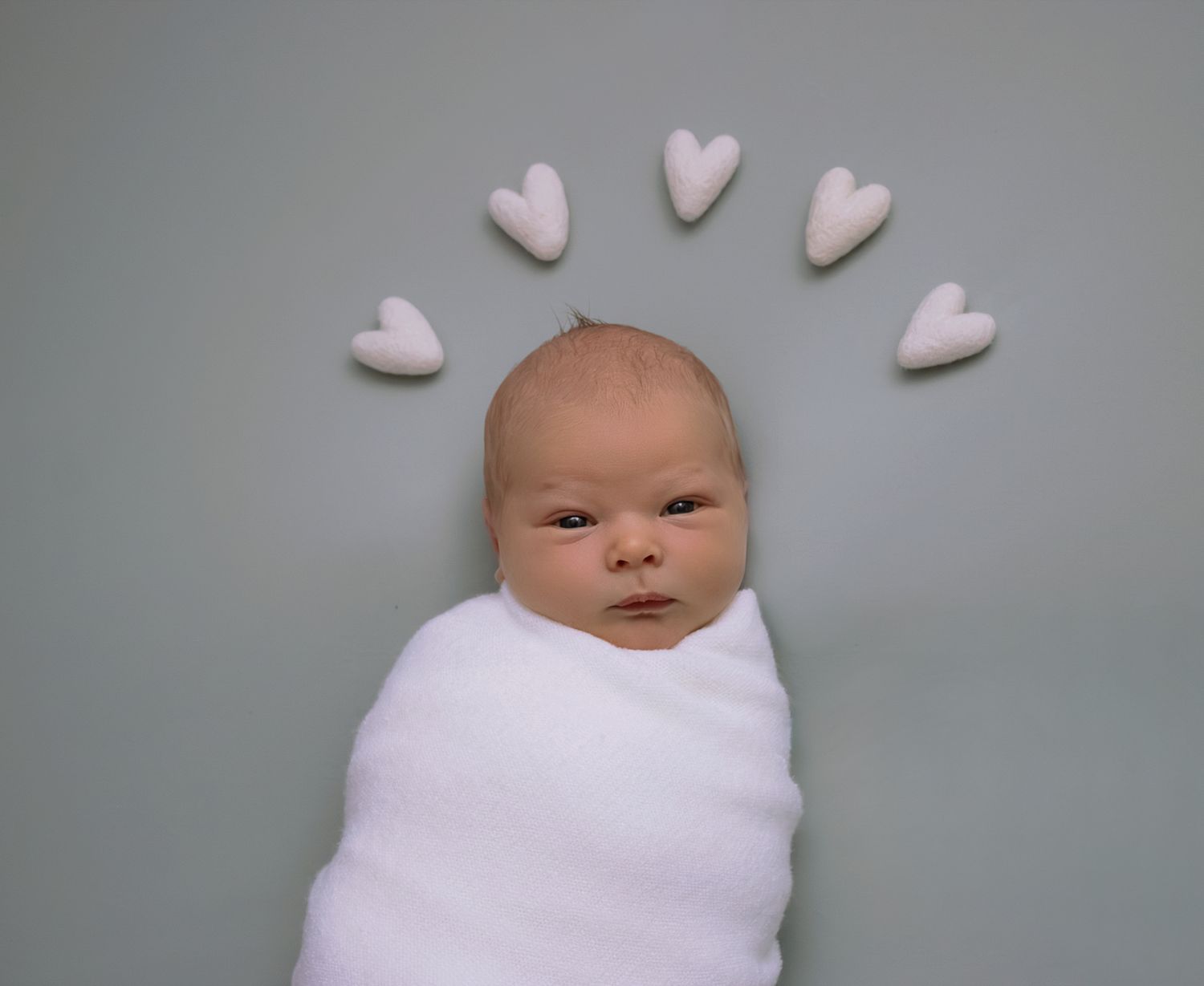Newborn baby swaddled in white wrap lying on gray background surrounded by small white heart shapes arranged in an arc.