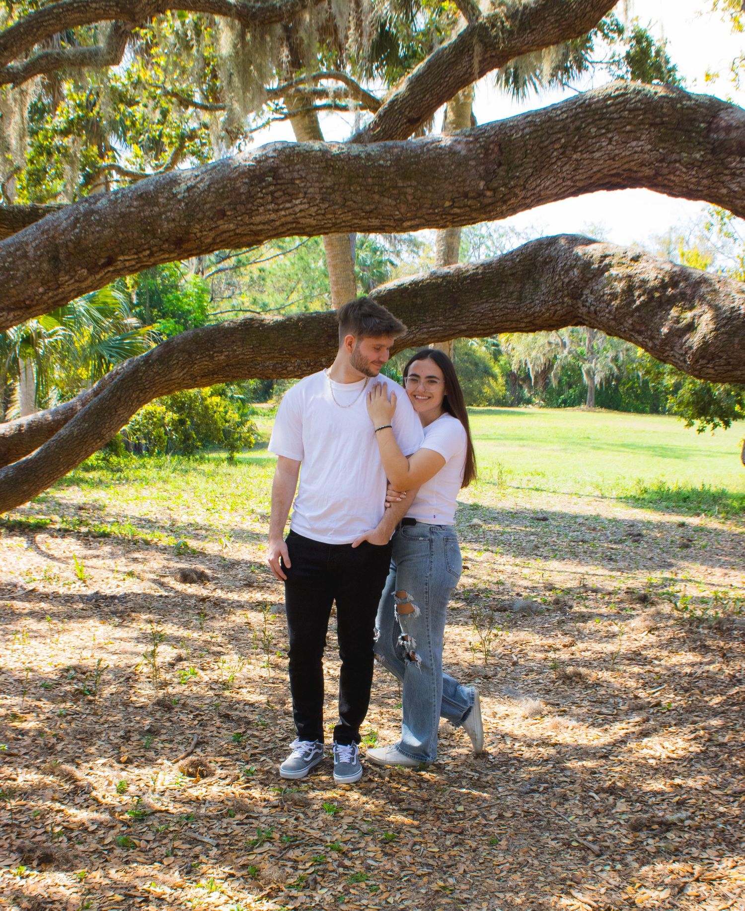 Couple posing under a large oak tree in a sunny park, wearing white shirts and casual attire.