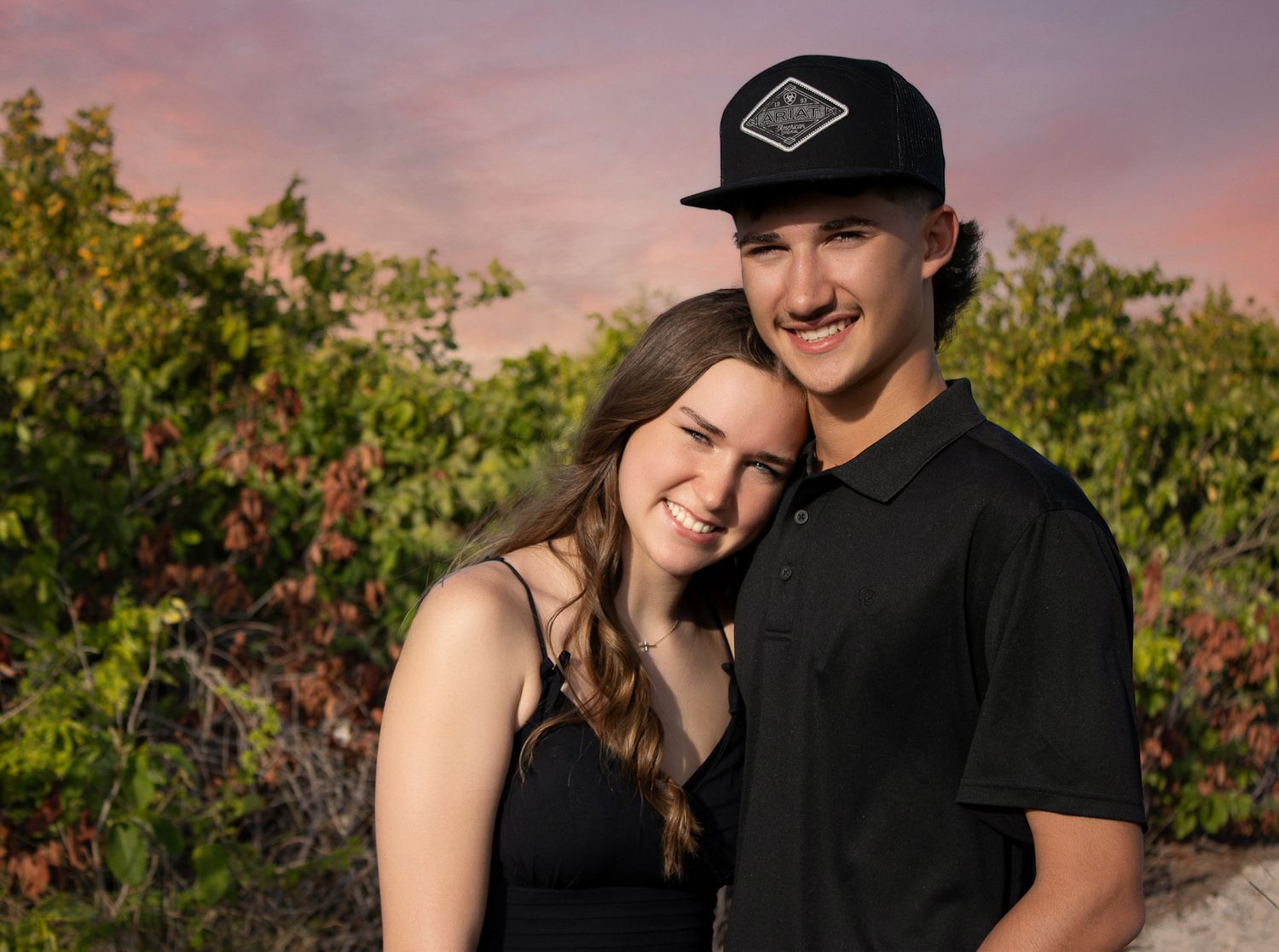 A smiling young couple poses outdoors at sunset, woman in black top leaning on man wearing black polo and snapback hat.
