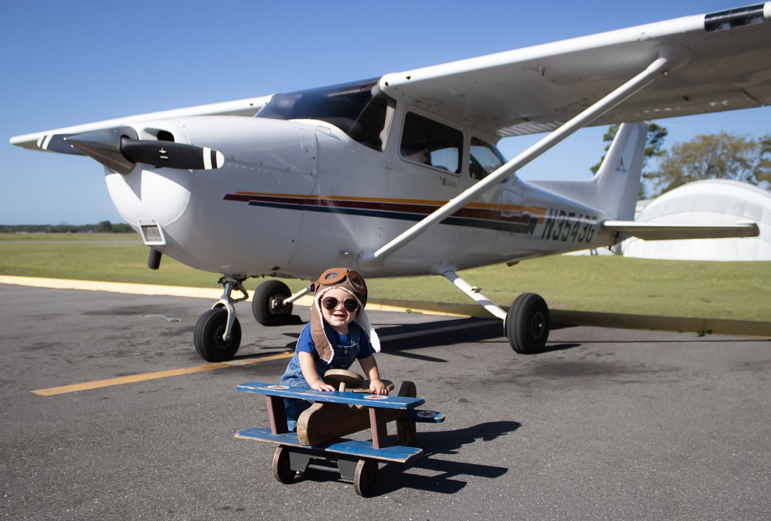 Young child riding a toy airplane on a tarmac in front of a white Cessna aircraft on a sunny day.
