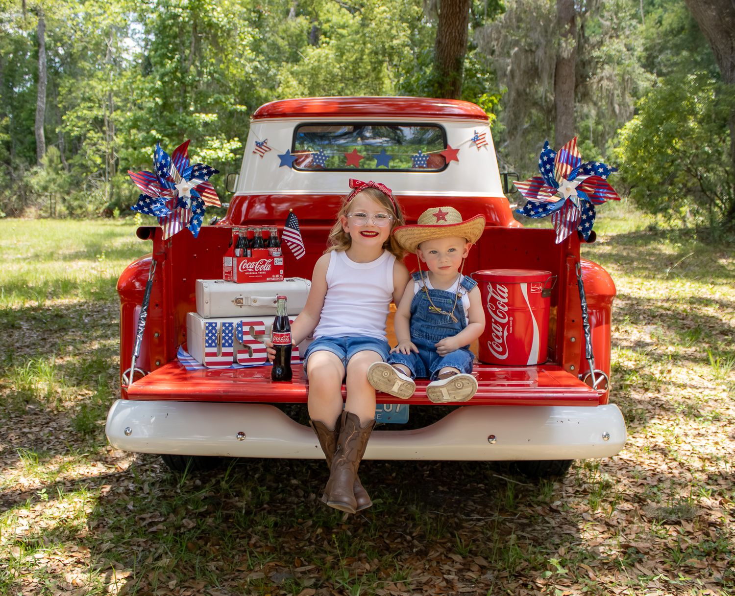 Two children sit in a red vintage truck bed with Coca-Cola coolers and patriotic pinwheels on a sunny day.
