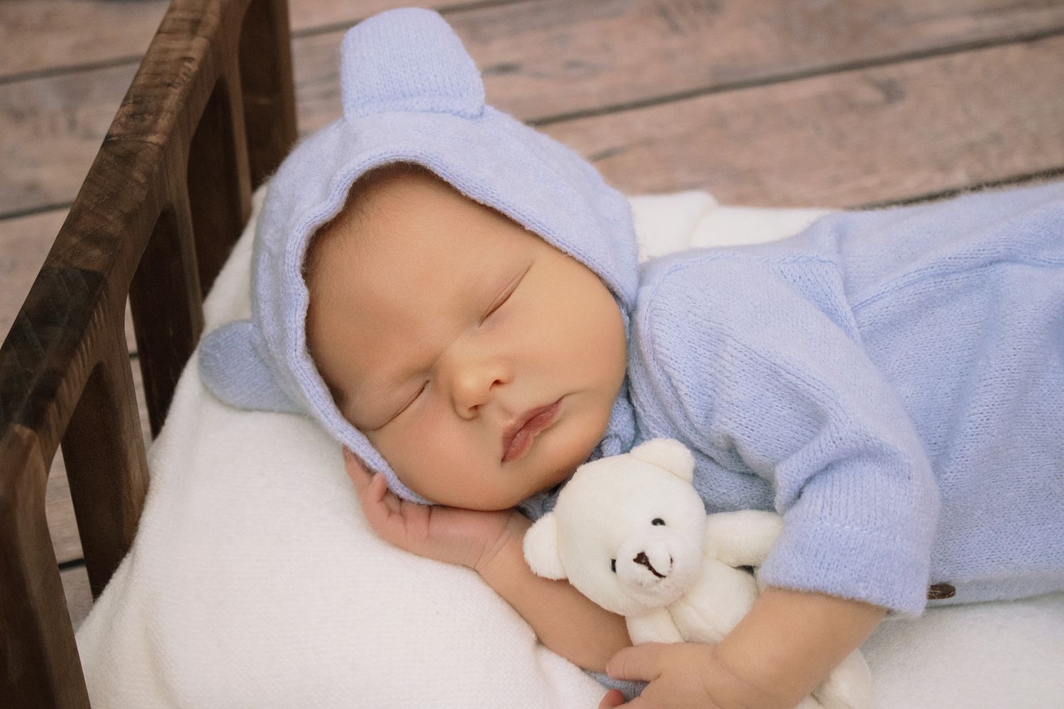 Sleeping newborn baby in blue bear onesie hugging a white teddy bear in a wooden crib.