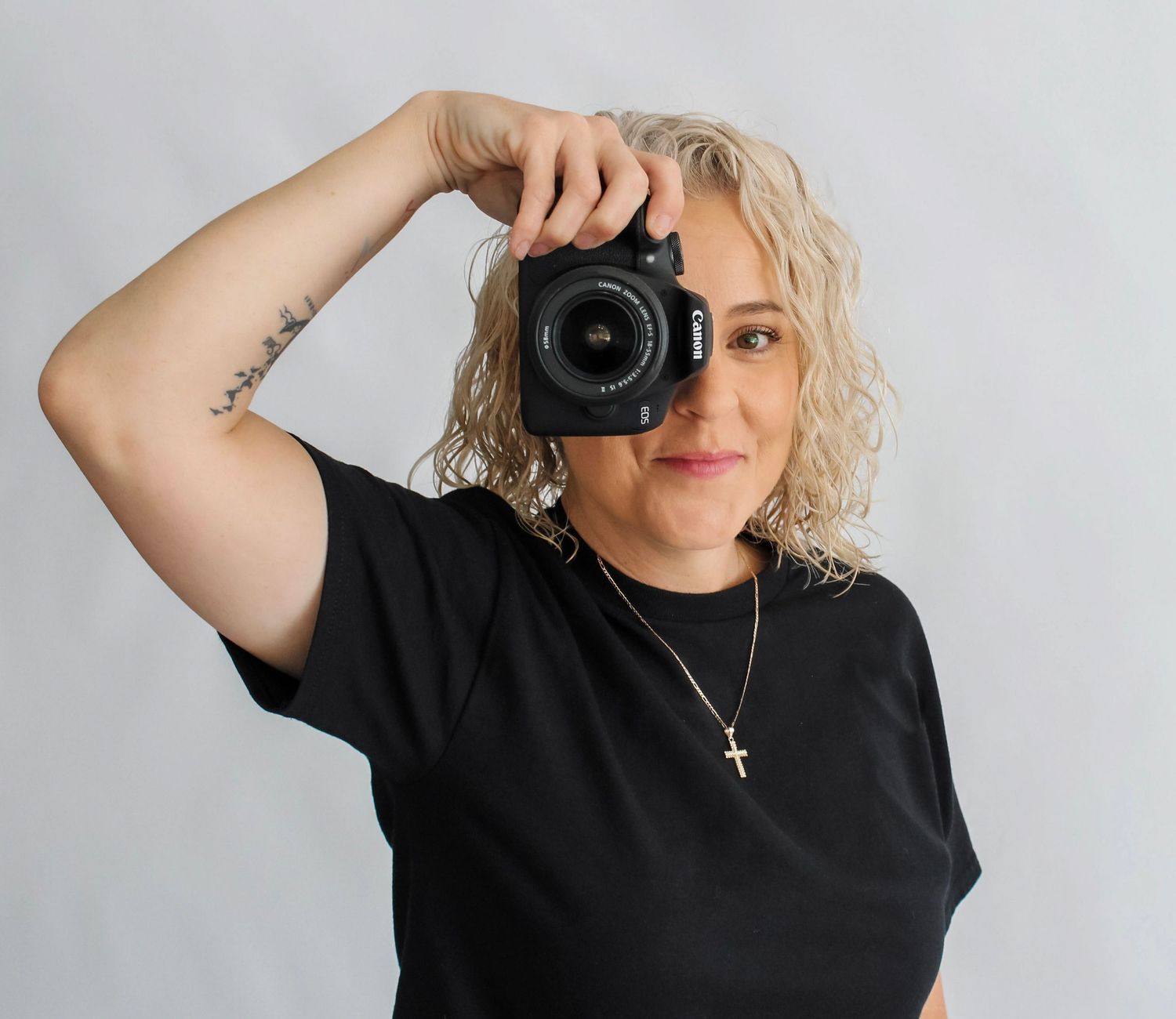 Woman in black t-shirt holding Canon DSLR camera up to her face, wearing a gold cross necklace, curly blonde hair.