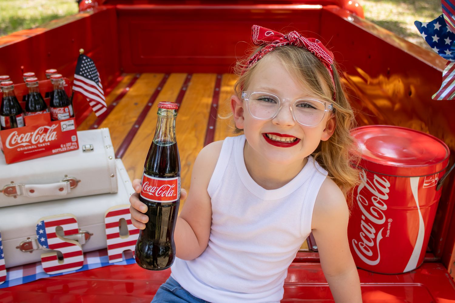 Young girl with red bow headband holding Coca-Cola bottle in festive truck bed with American flags.