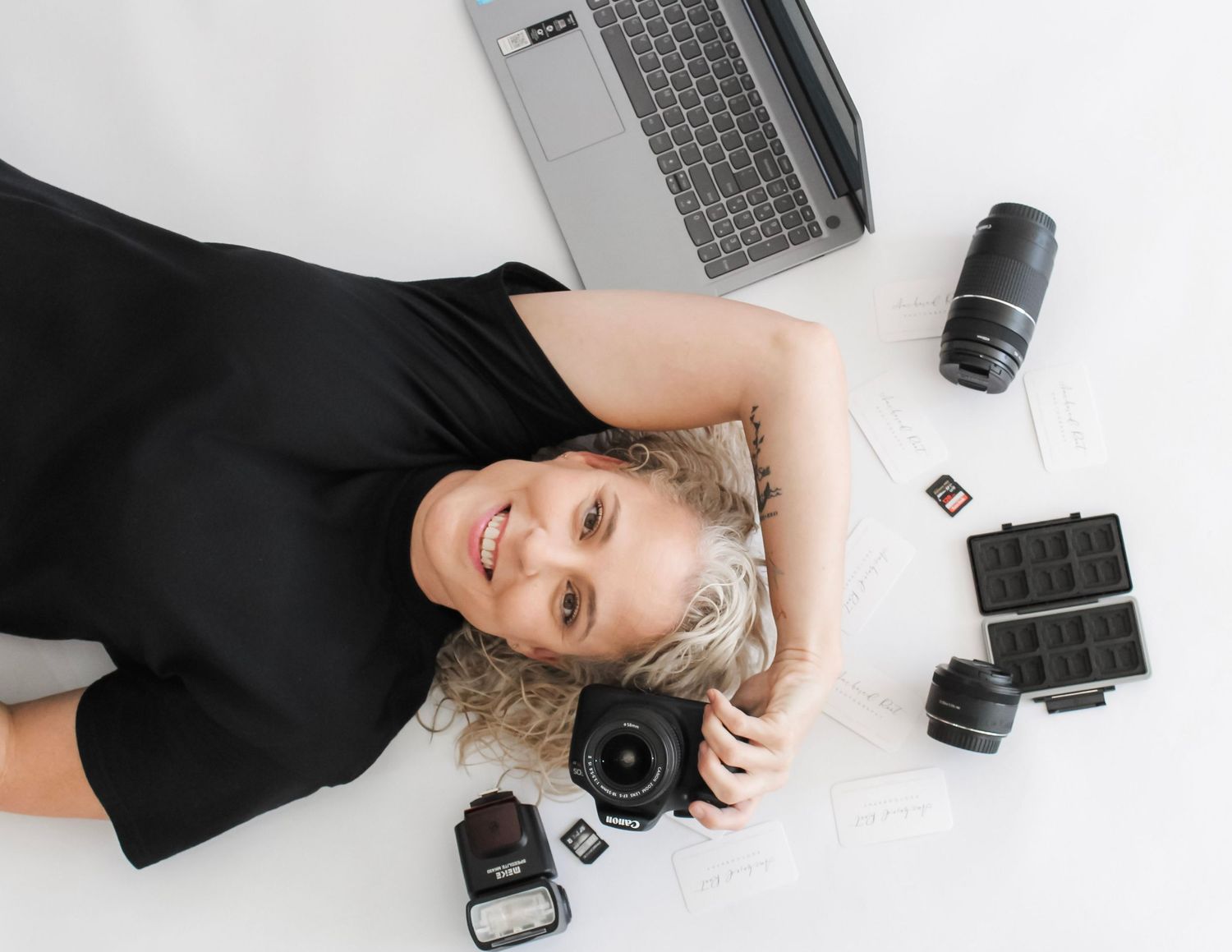 A smiling female photographer lying on a white floor surrounded by camera equipment, lenses, memory cards, and a laptop.
