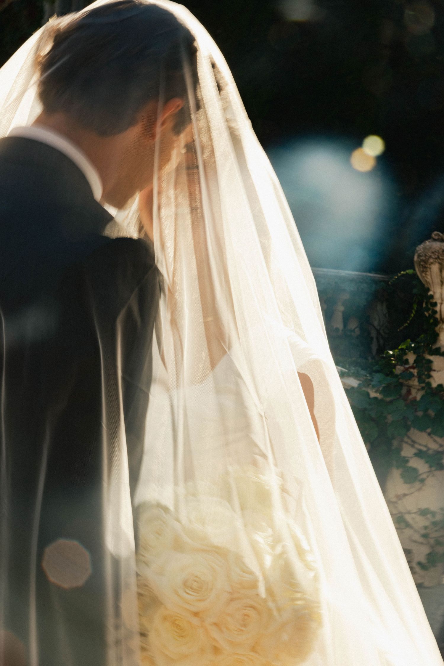 Bride and groom share romantic kiss under flowing wedding veil, bride holding white rose bouquet in soft golden light.
