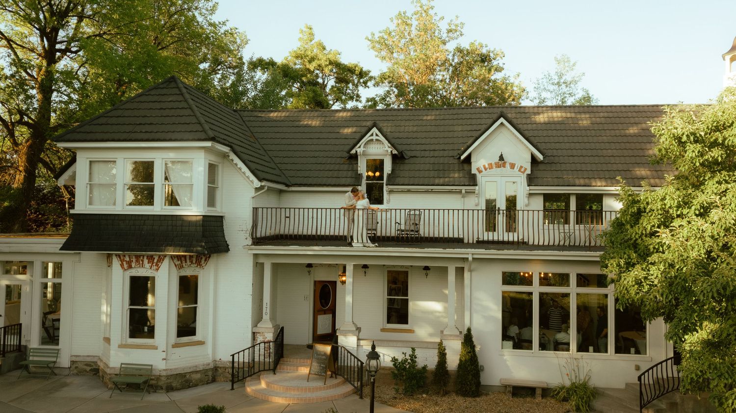 Elegant white Victorian-style house with wraparound porch and balcony set among green trees.