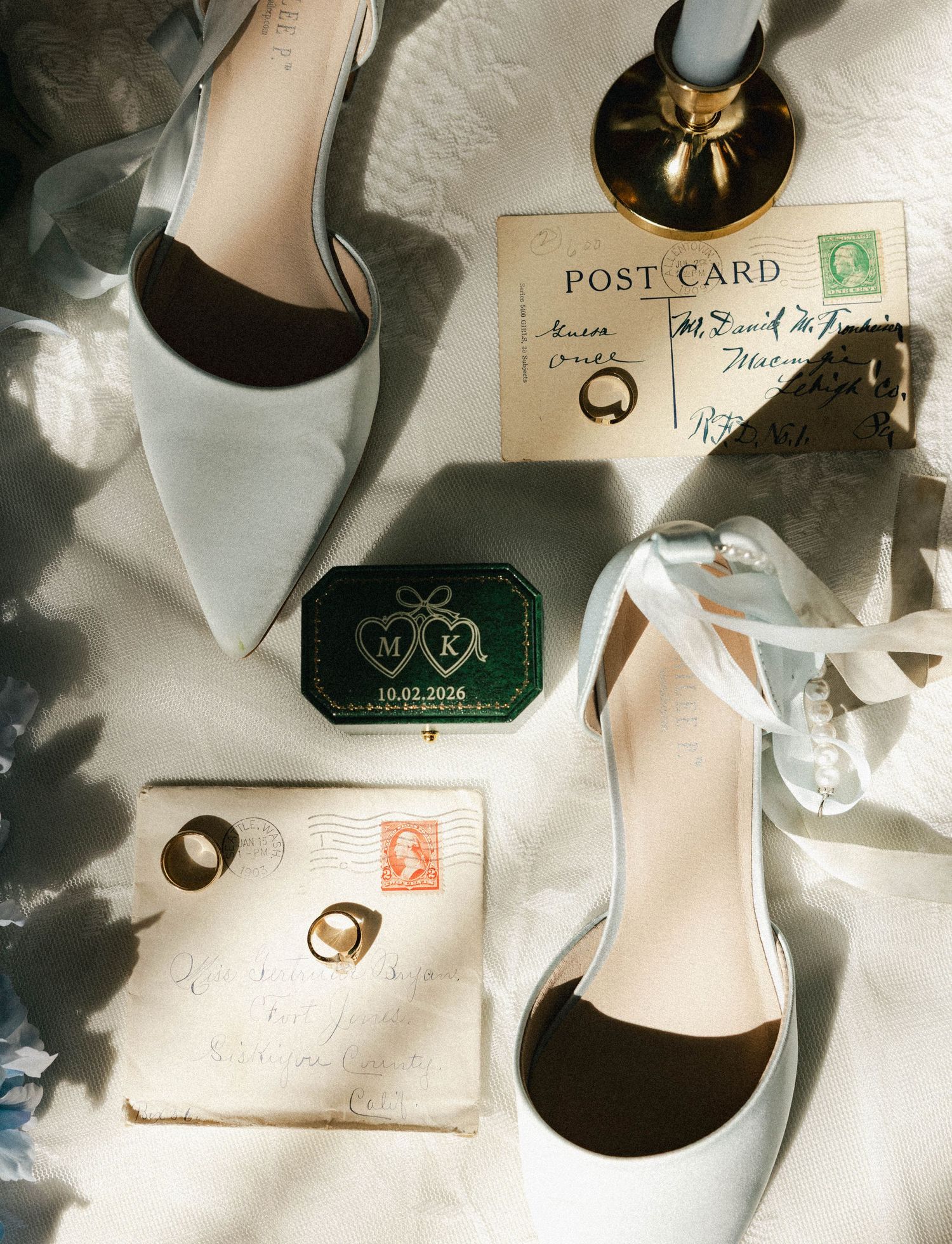 Elegant wedding flat lay featuring white satin heels, velvet ring box, vintage postcards, gold rings, and a brass candlestick.