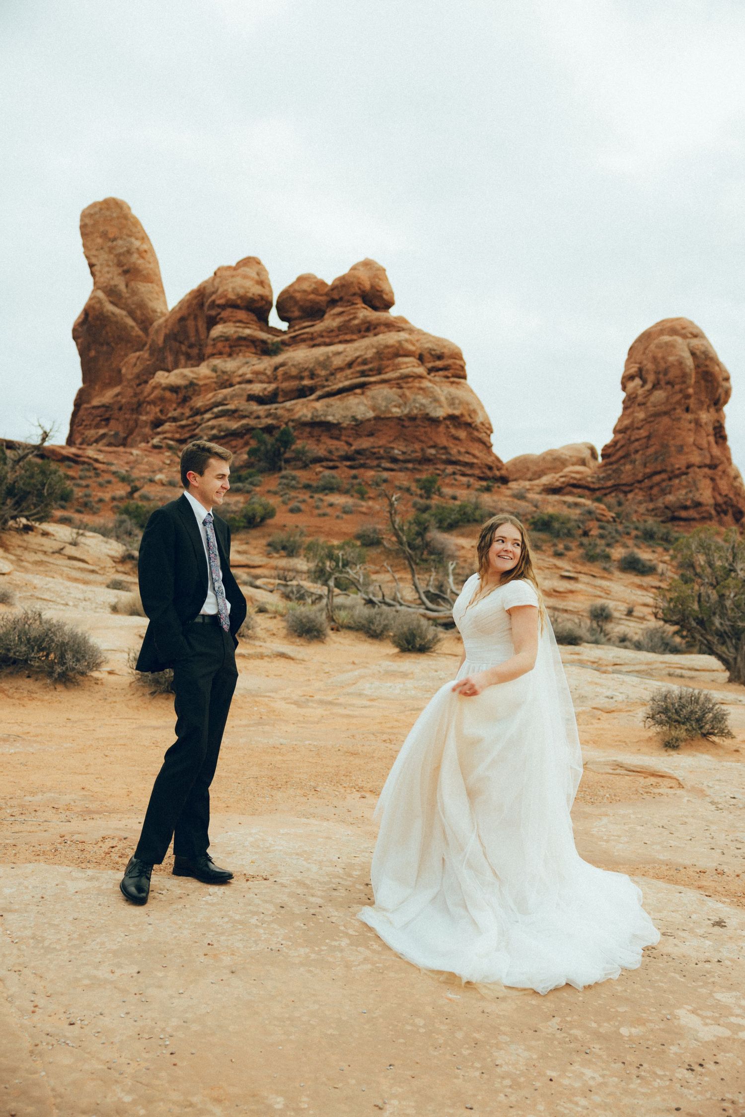 A couple in wedding attire dances and twirls against a backdrop of red rock formations in a desert landscape.