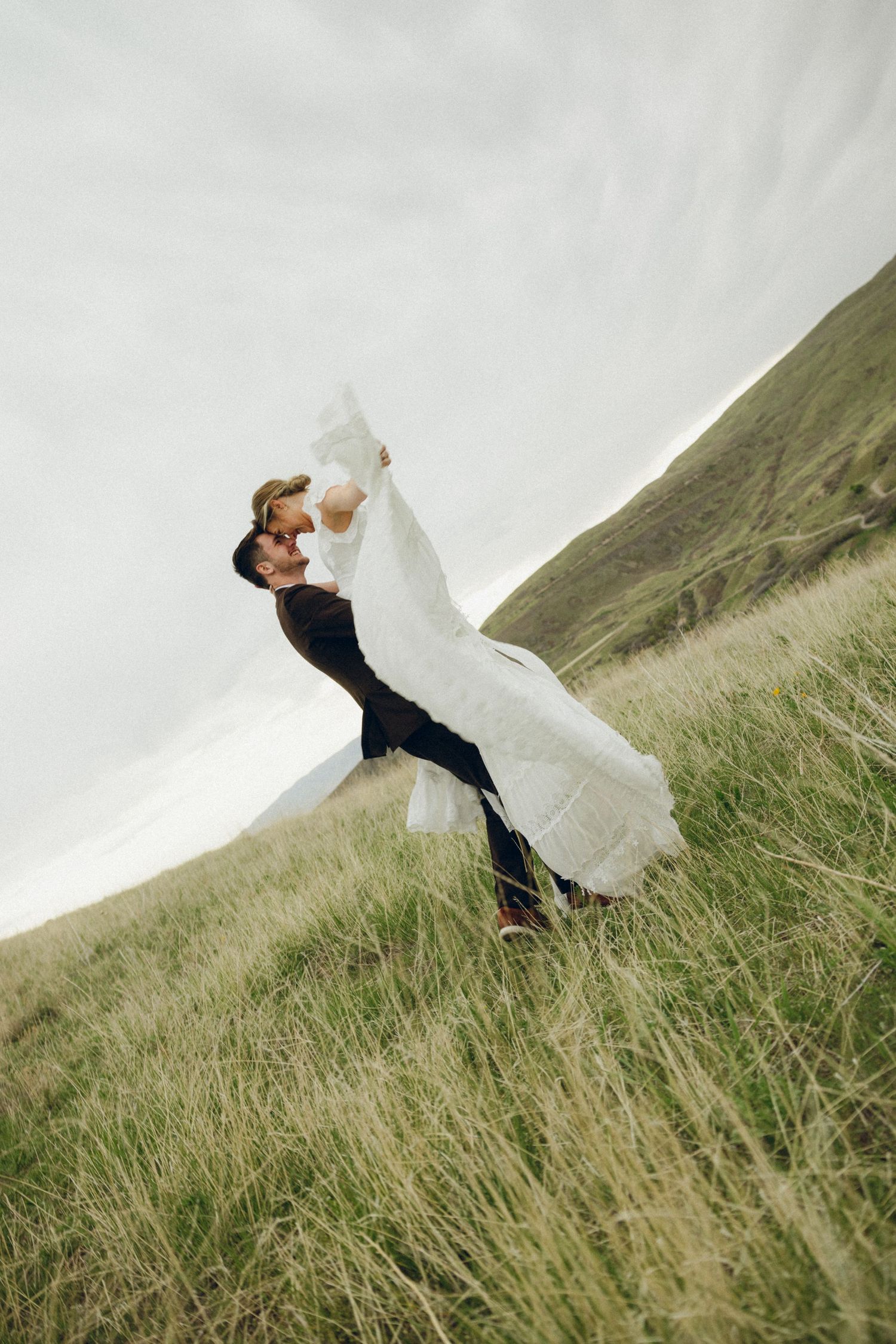 A couple celebrates their wedding day on a windswept grassy hillside with dramatic movement of the bride's flowing white dress.