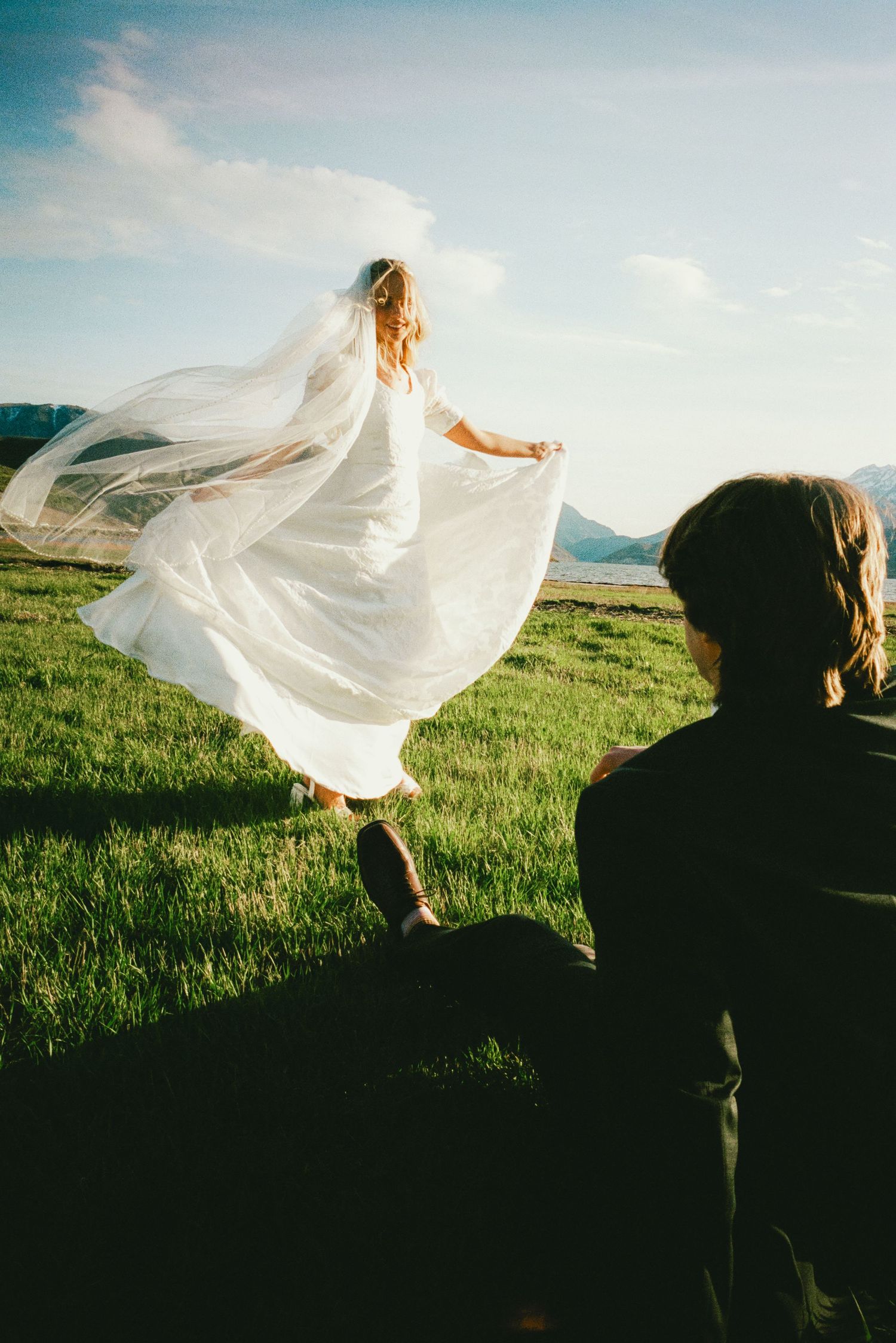 Bride in flowing white gown and veil dancing in green meadow while groom watches, mountains in background.
