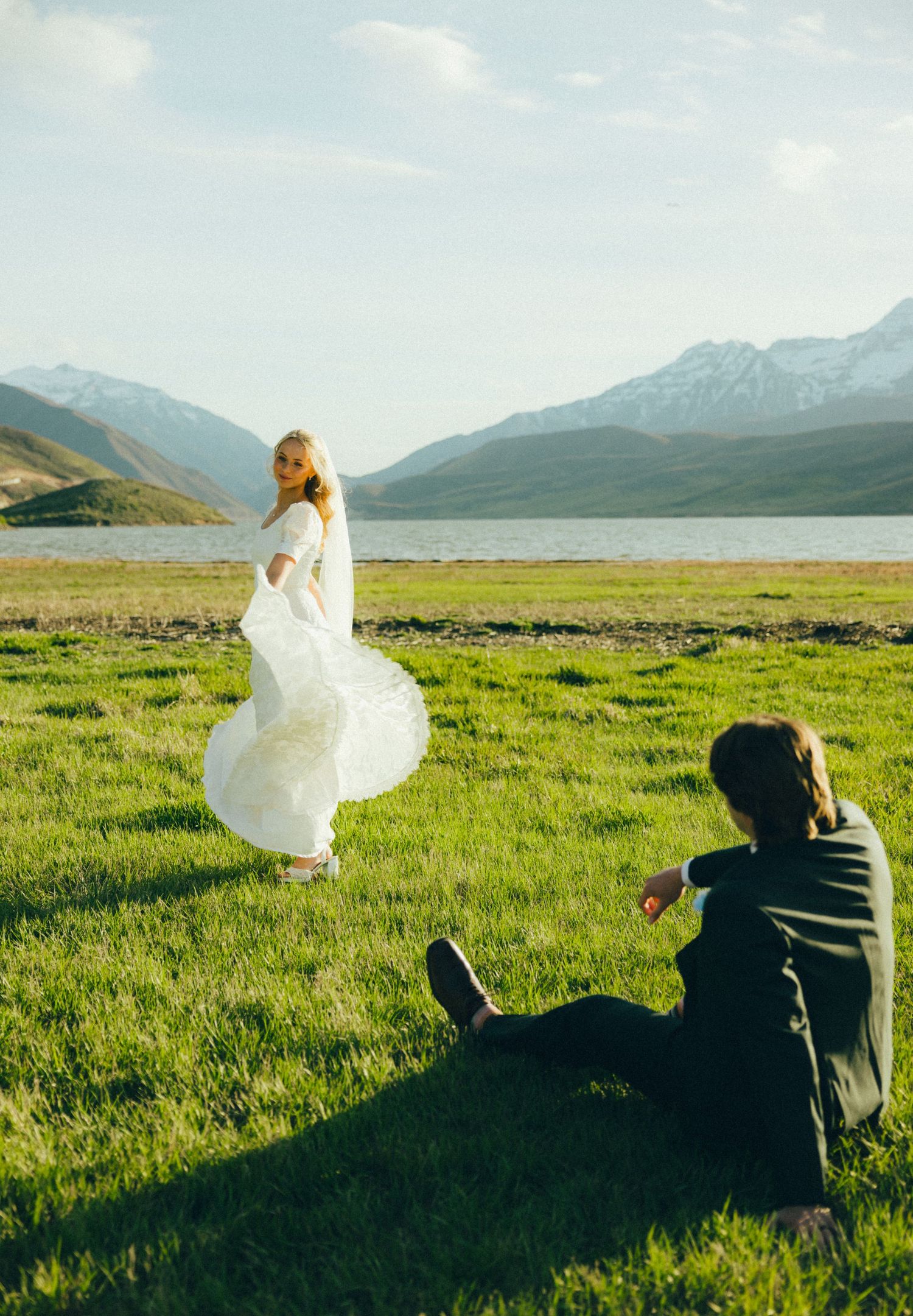 Bride in white gown twirling by a mountain lake while groom sits on green grass watching her.
