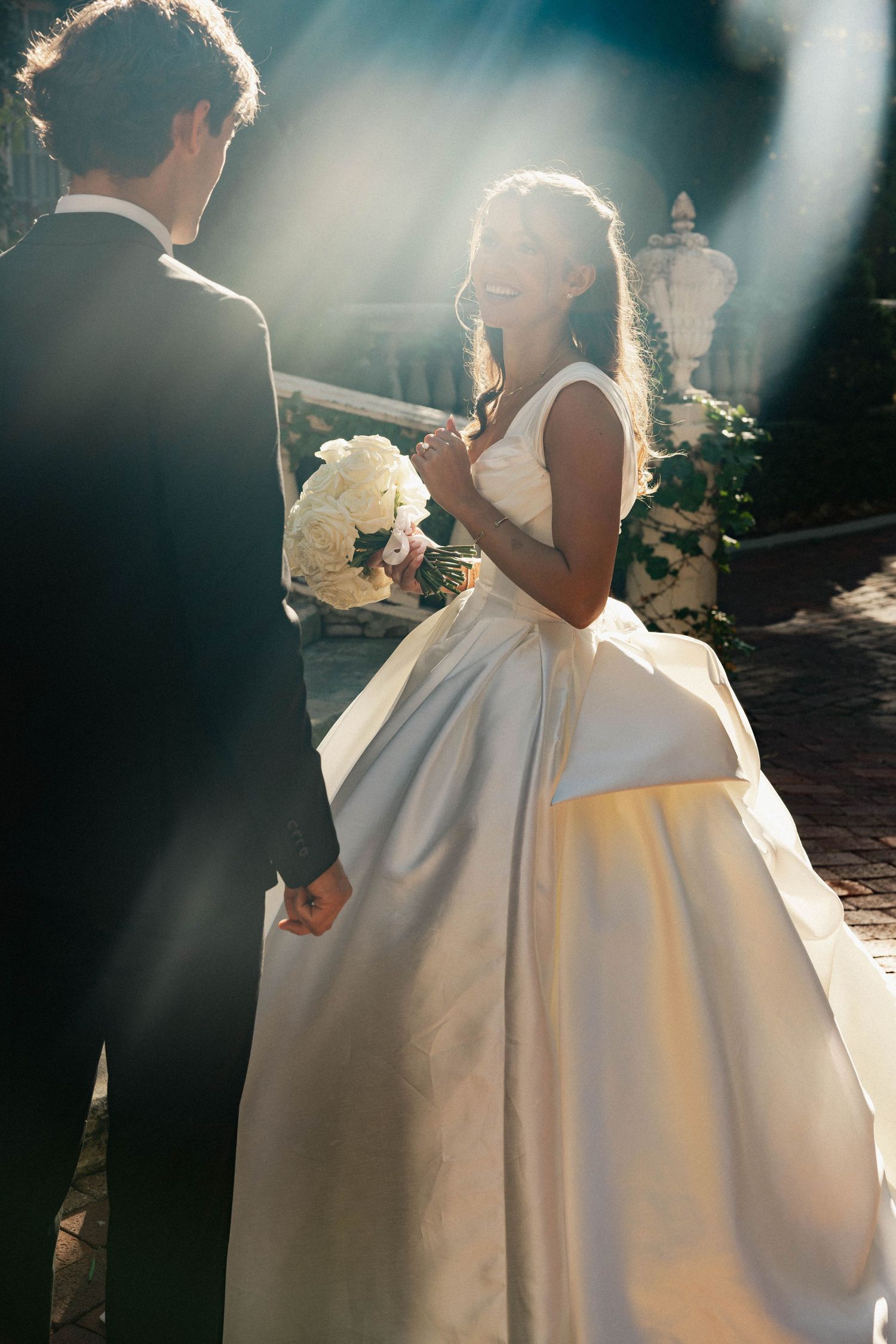 Bride in white ball gown holds bouquet while groom in black suit holds her hand in sunlit outdoor wedding.