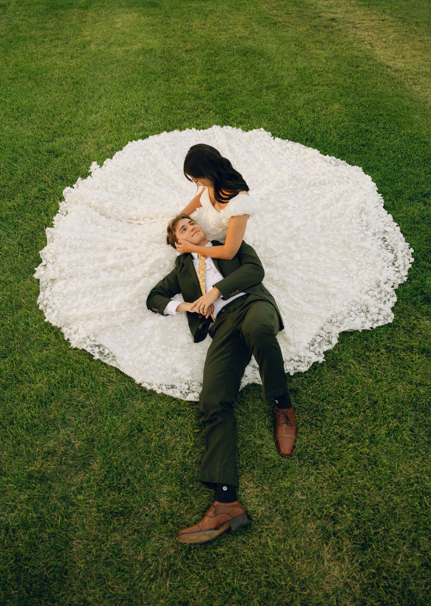 A couple in wedding attire embraces while lying on lush green grass, with the bride's white dress spread out beautifully.