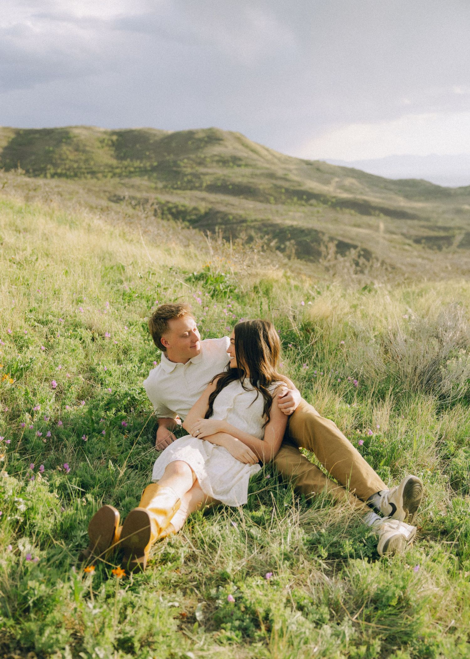 Couple sitting together on a lush green hillside, sharing a romantic moment with rolling mountains in the background.