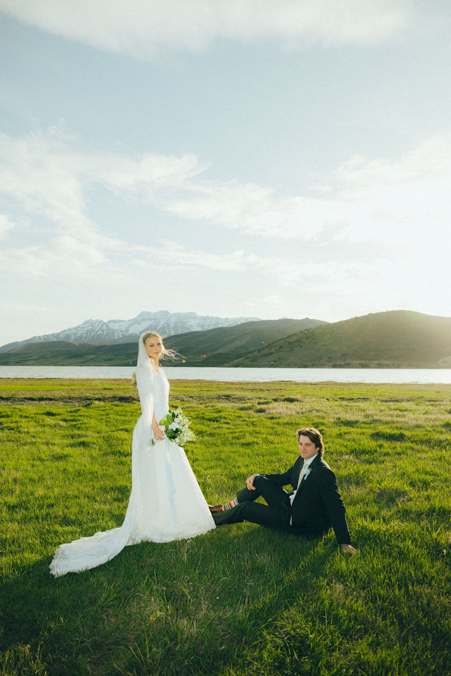 Bride in white gown and groom in black suit pose in green meadow with mountain lake backdrop on wedding day.