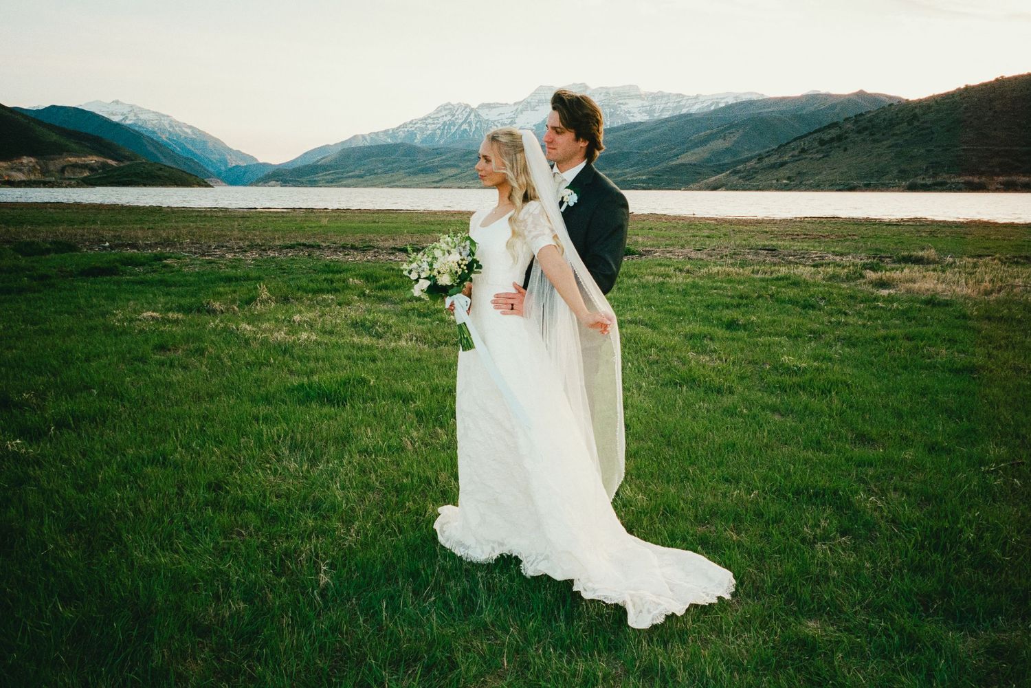 Bride and groom stand in lush green field with snow-capped mountains and lake backdrop at outdoor wedding.