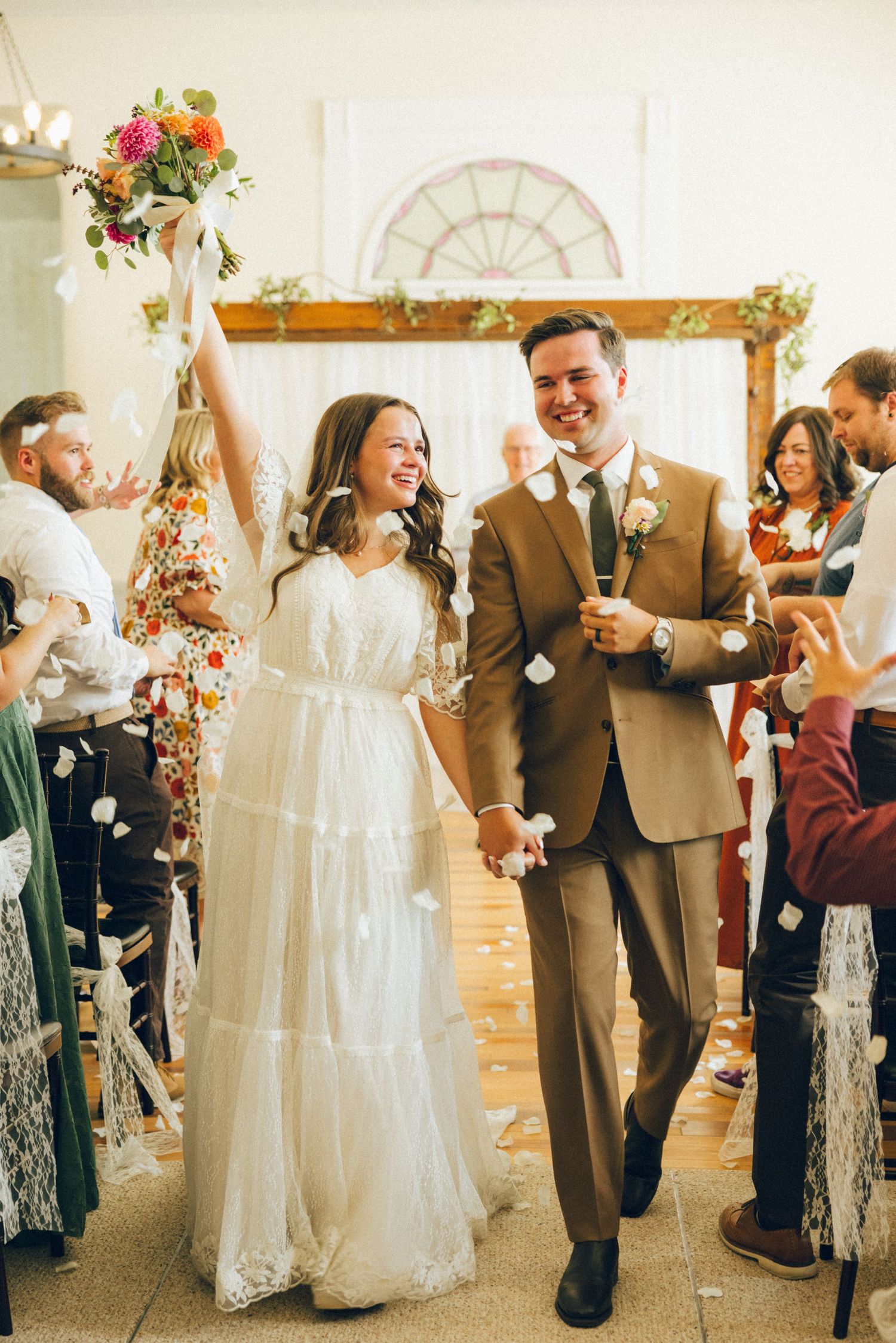 Wedding guests celebrate and dance in a bright indoor venue with white lace and floral decorations.