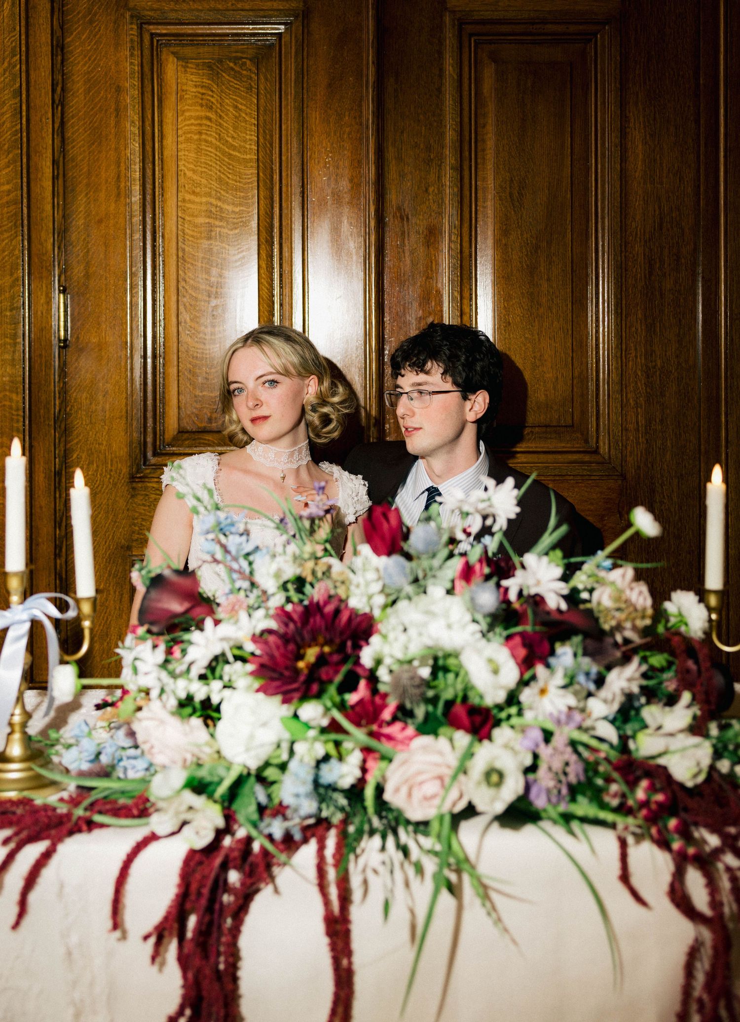 Elegant wedding couple seated at a floral sweetheart table with burgundy and white blooms and candlelight in a wood-paneled room.