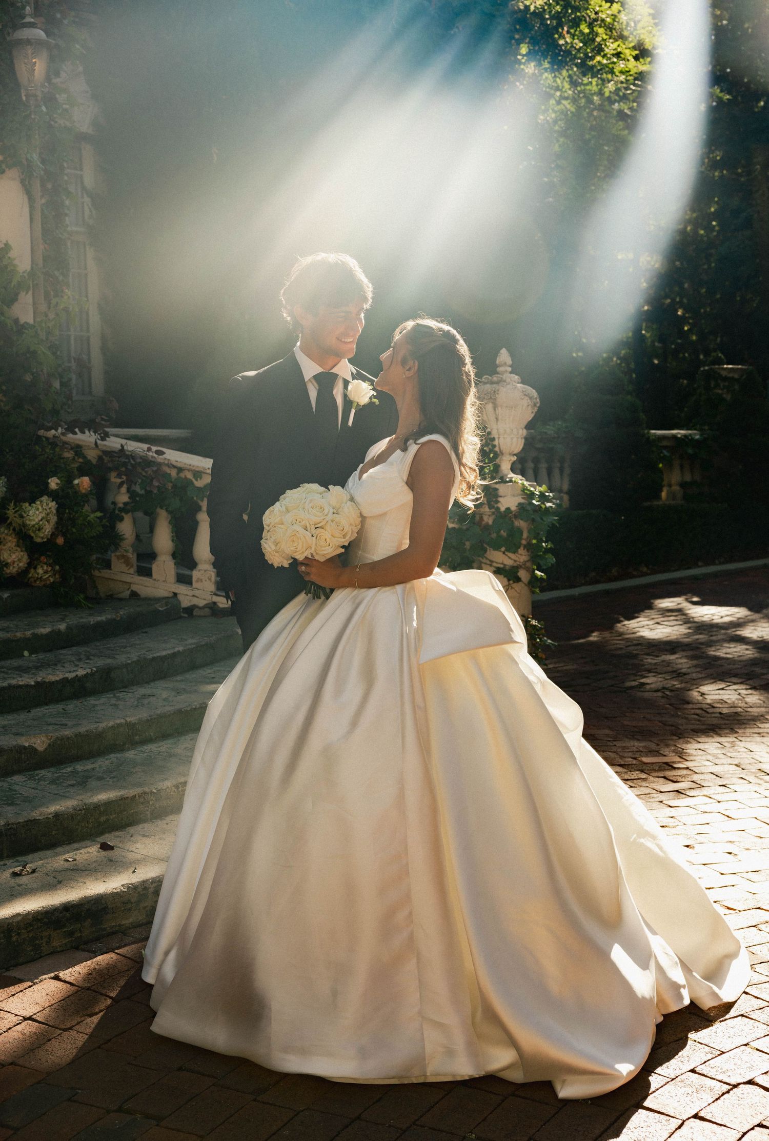 Bride and groom share a romantic moment in golden sunlight, she holds white bouquet in elegant ball gown at outdoor venue.