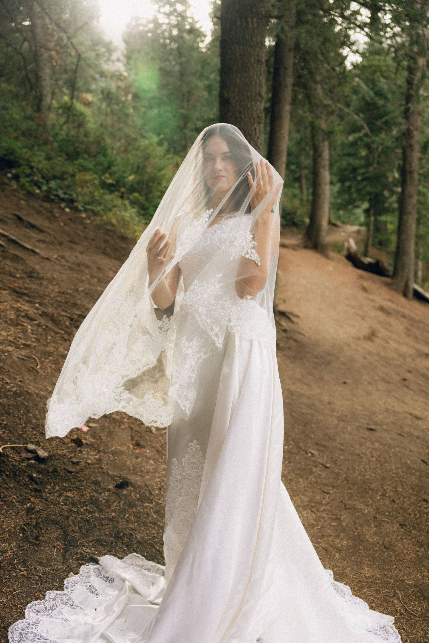 Elegant white lace wedding gown with long veil photographed on forest trail during dreamy outdoor bridal session.