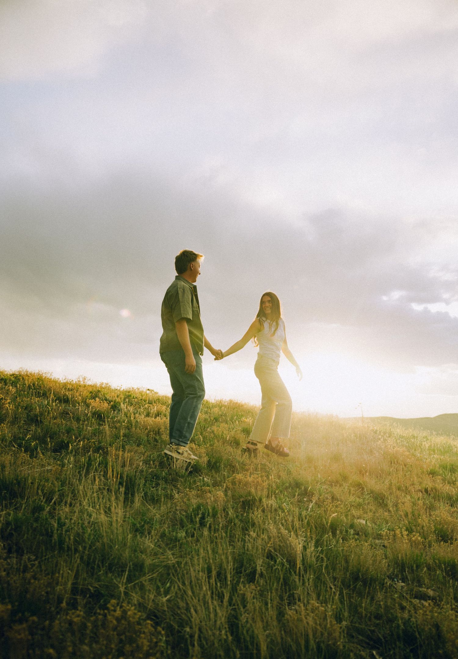 Couple holding hands on a sunlit grassy hill at golden hour with dramatic cloudy sky backdrop.