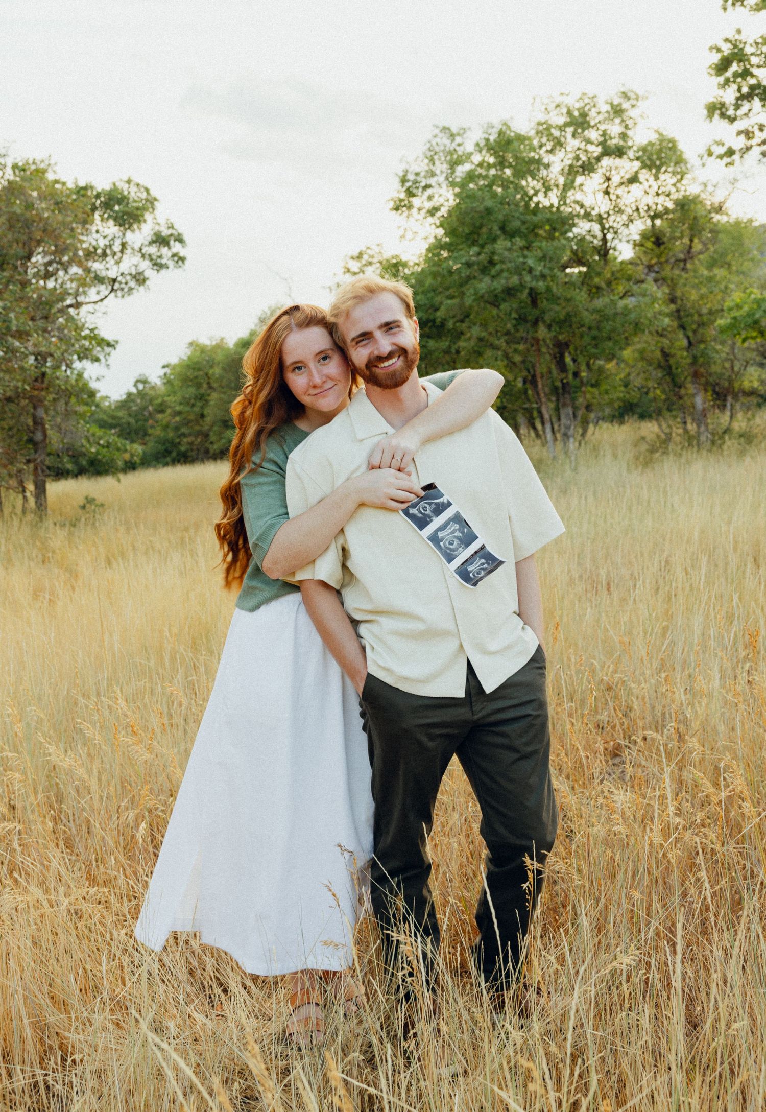A couple poses together in a sunny field of tall grass wearing light colored clothing.