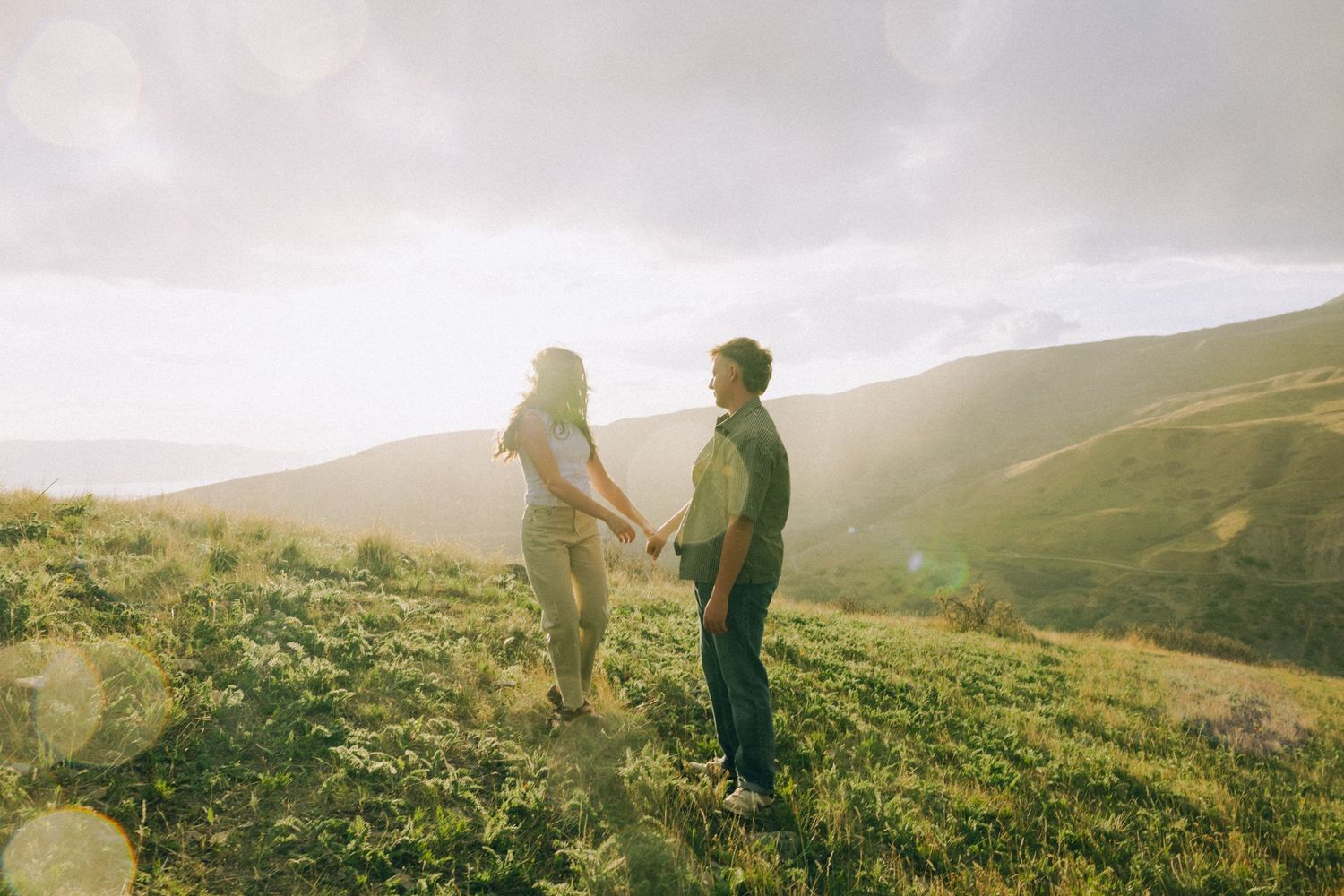 Couple holding hands on a lush green hillside with golden sunlight and rolling mountains in the background.