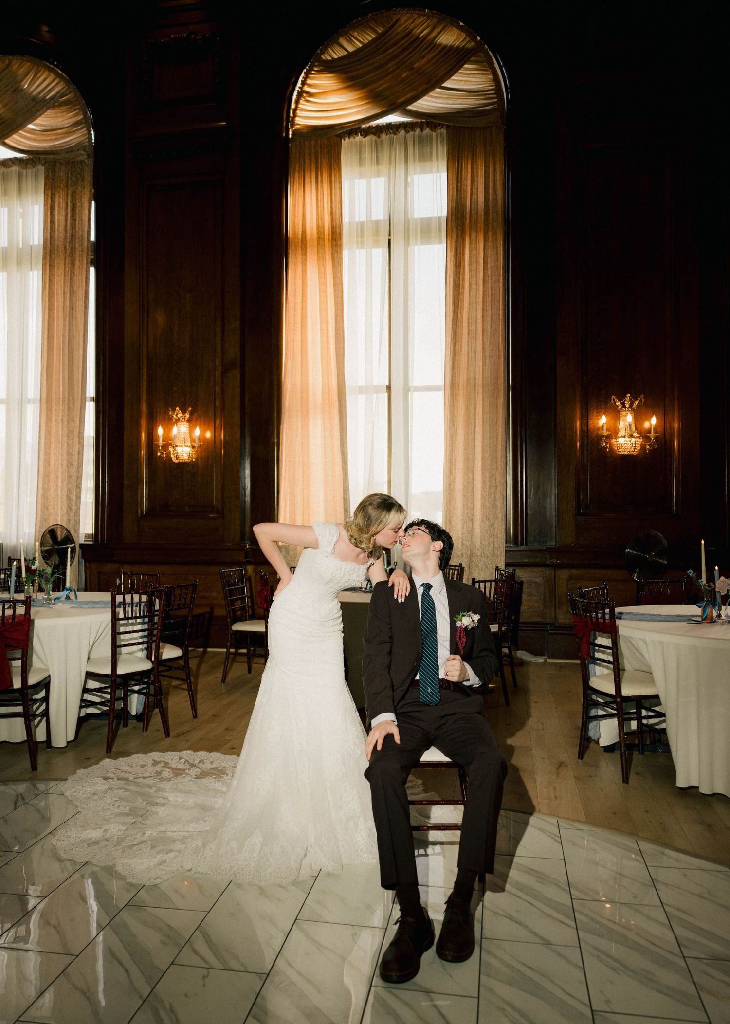 Bride in white gown kisses groom seated in chair at elegant wedding reception with wood paneling and draped windows.