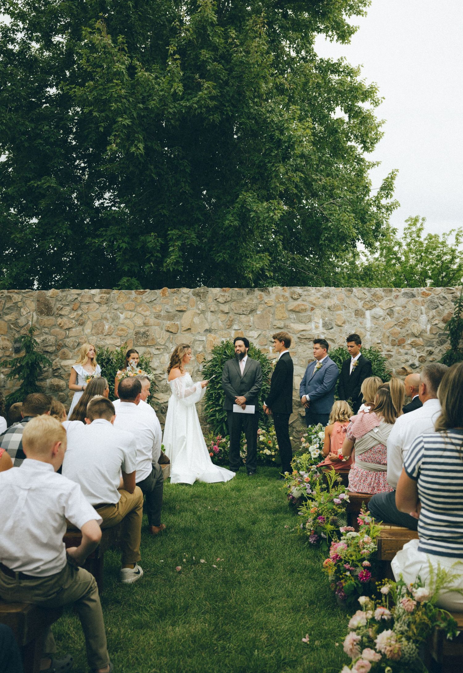 An outdoor wedding ceremony takes place in a garden setting with stone walls and lush greenery in the background.