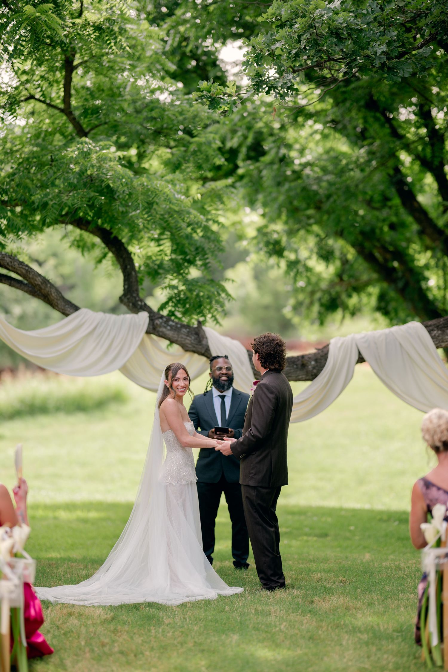 Outdoor wedding ceremony under large tree with white draping, bride and groom exchanging vows with officiant.