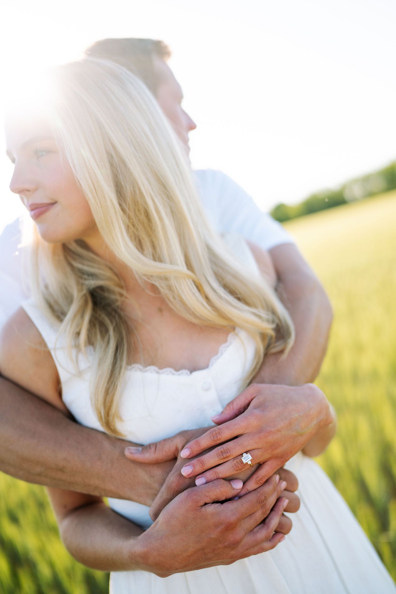 Couple embracing in a sunny field, woman wearing an engagement ring, both dressed in white.
