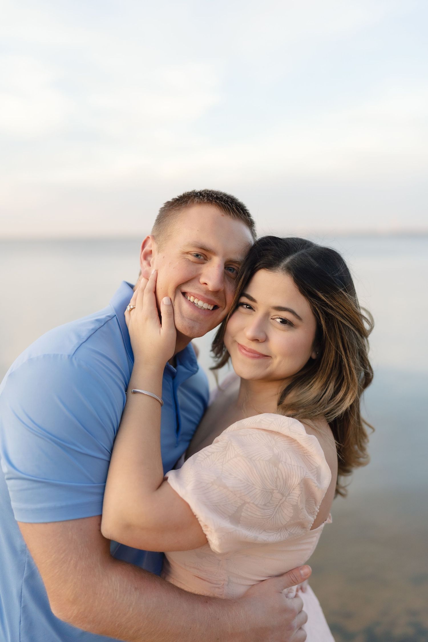 A couple shares an intimate embrace against a soft cloudy sky background during a beach photoshoot.