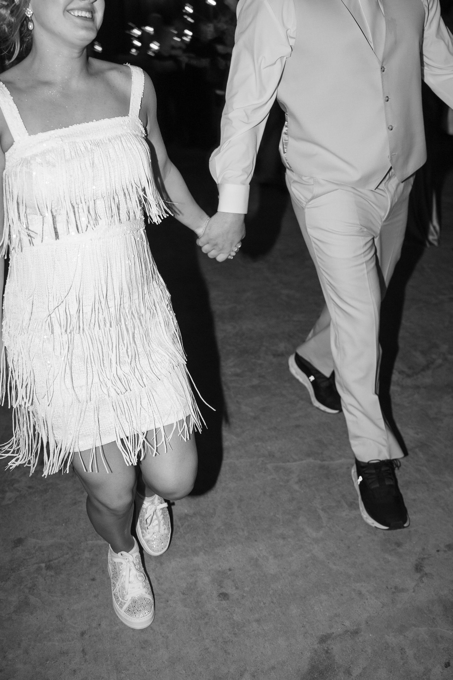 Wedding party dances in white attire with fringe details on a dark dance floor.