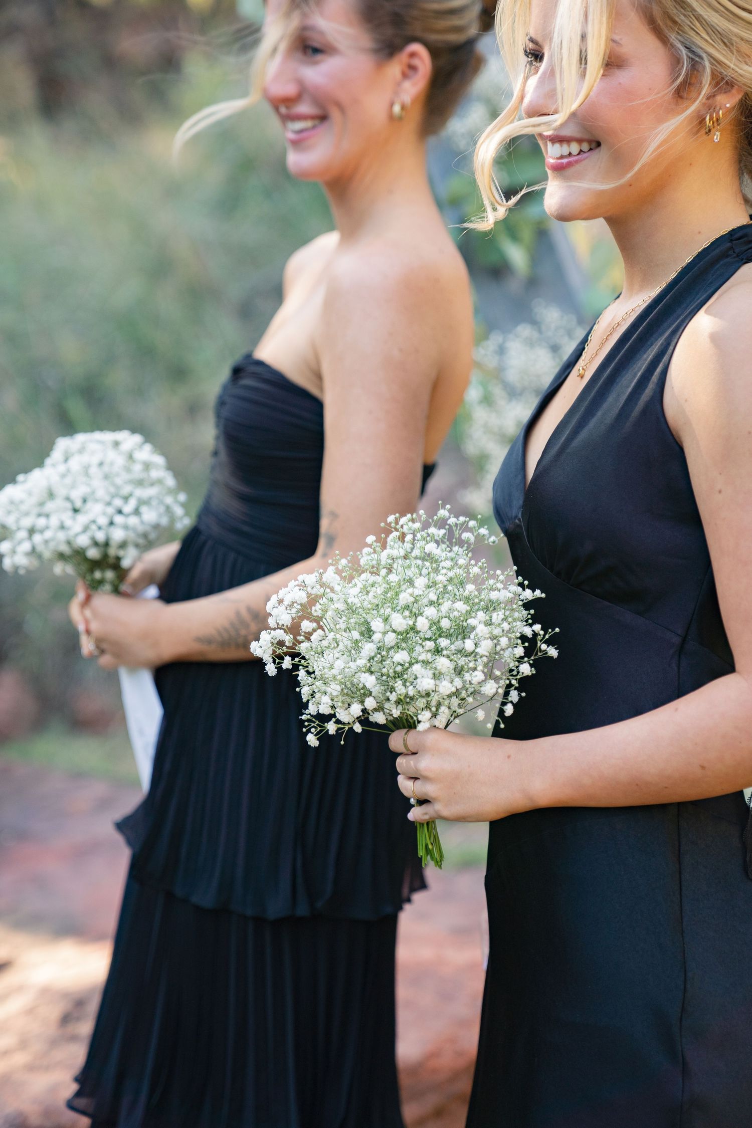 Bridesmaids in black dresses holding baby's breath bouquets stand together outdoors.