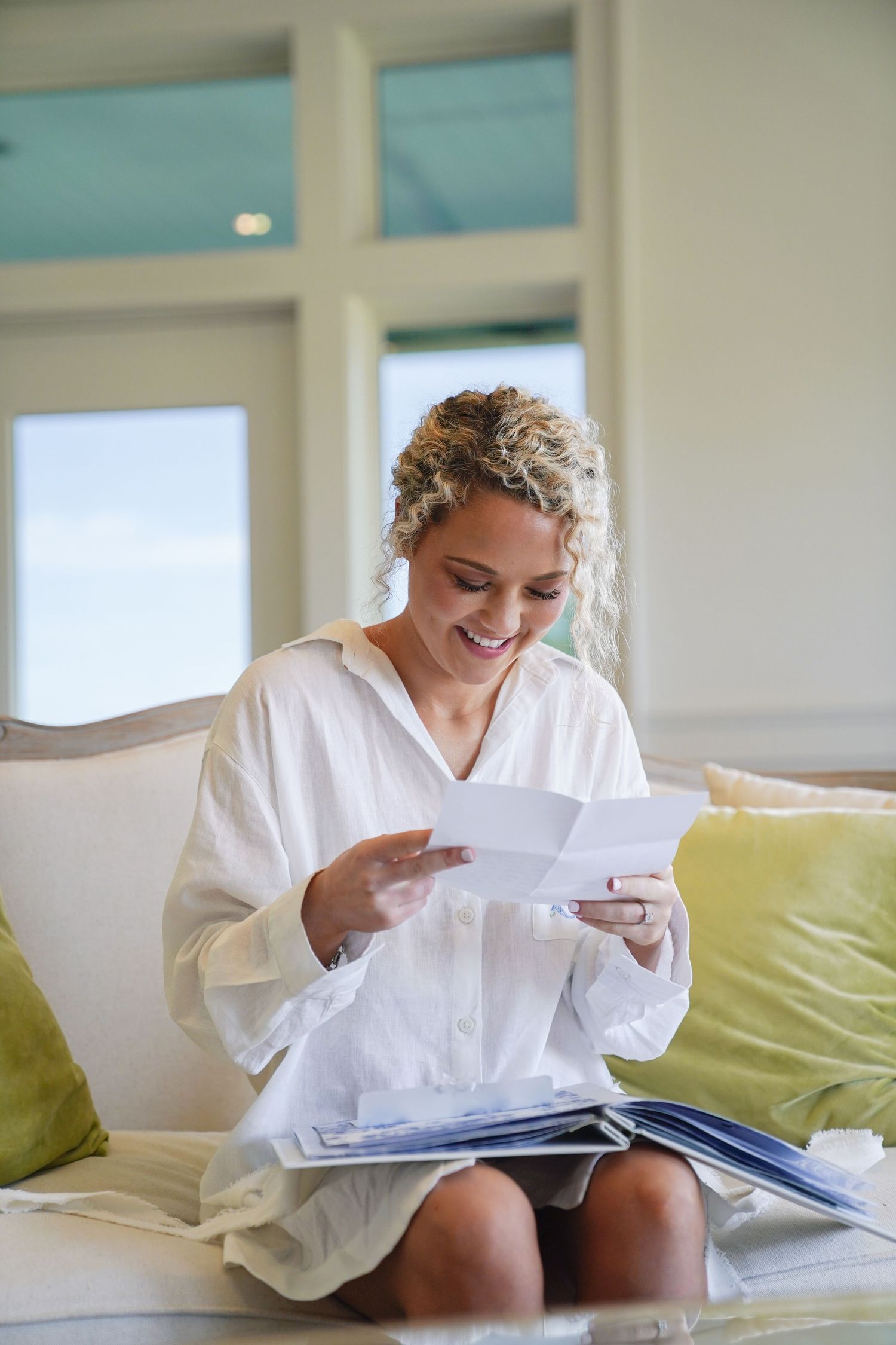 Someone in a white robe reads letters while sitting on a couch in a sunlit room.