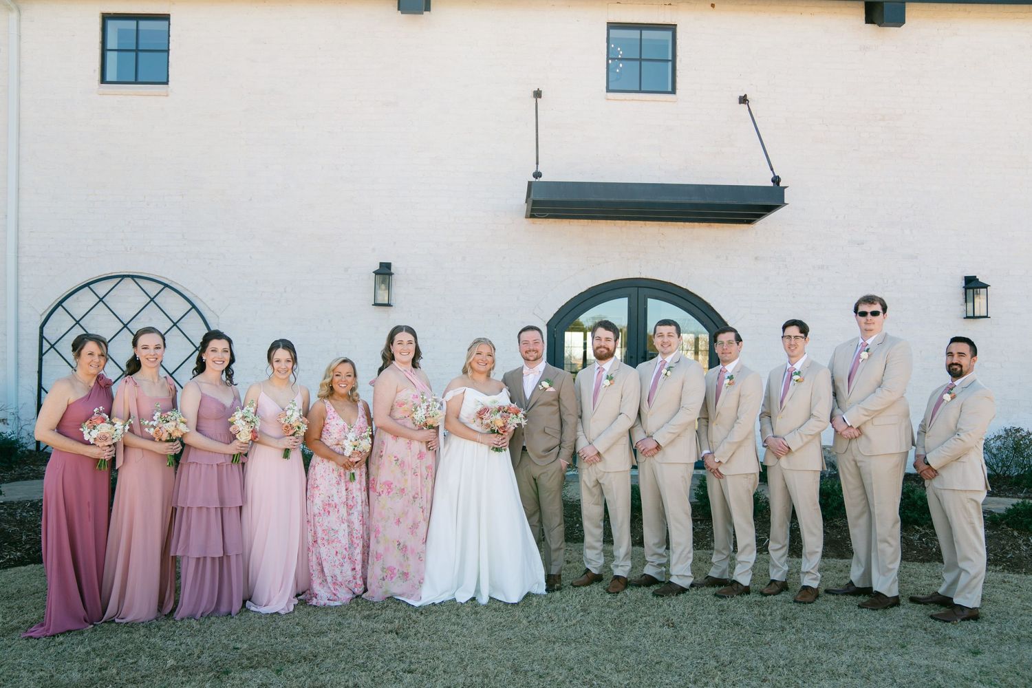 Wedding party lined up against white building wall for formal group photo.