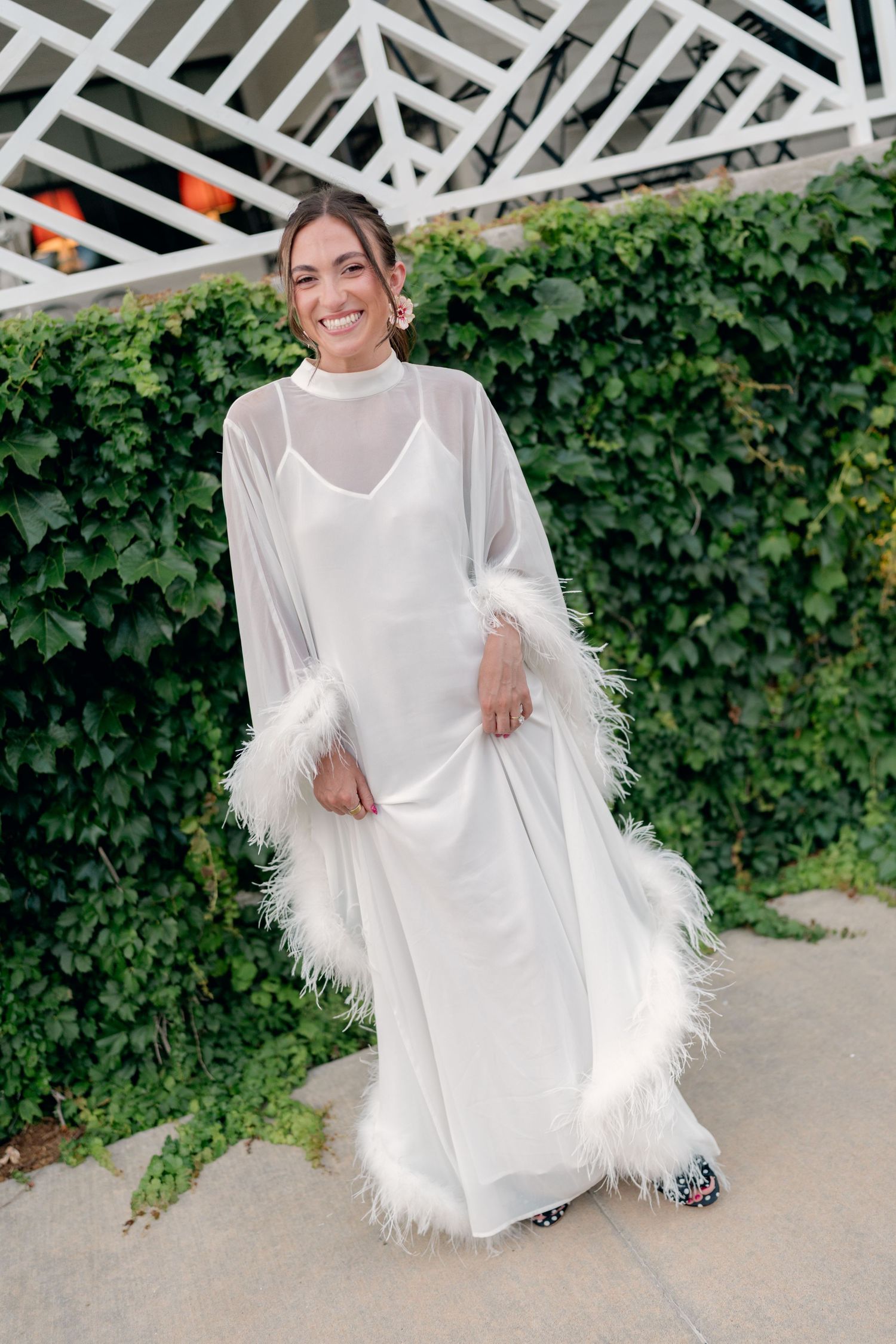 Woman in white feather-trimmed sheer wrap over satin slip dress, smiling outdoors against green ivy wall.