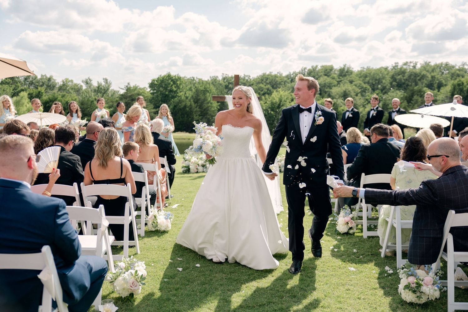 Bride and groom walk down outdoor aisle after wedding ceremony, guests seated on white chairs, lush green lawn venue.