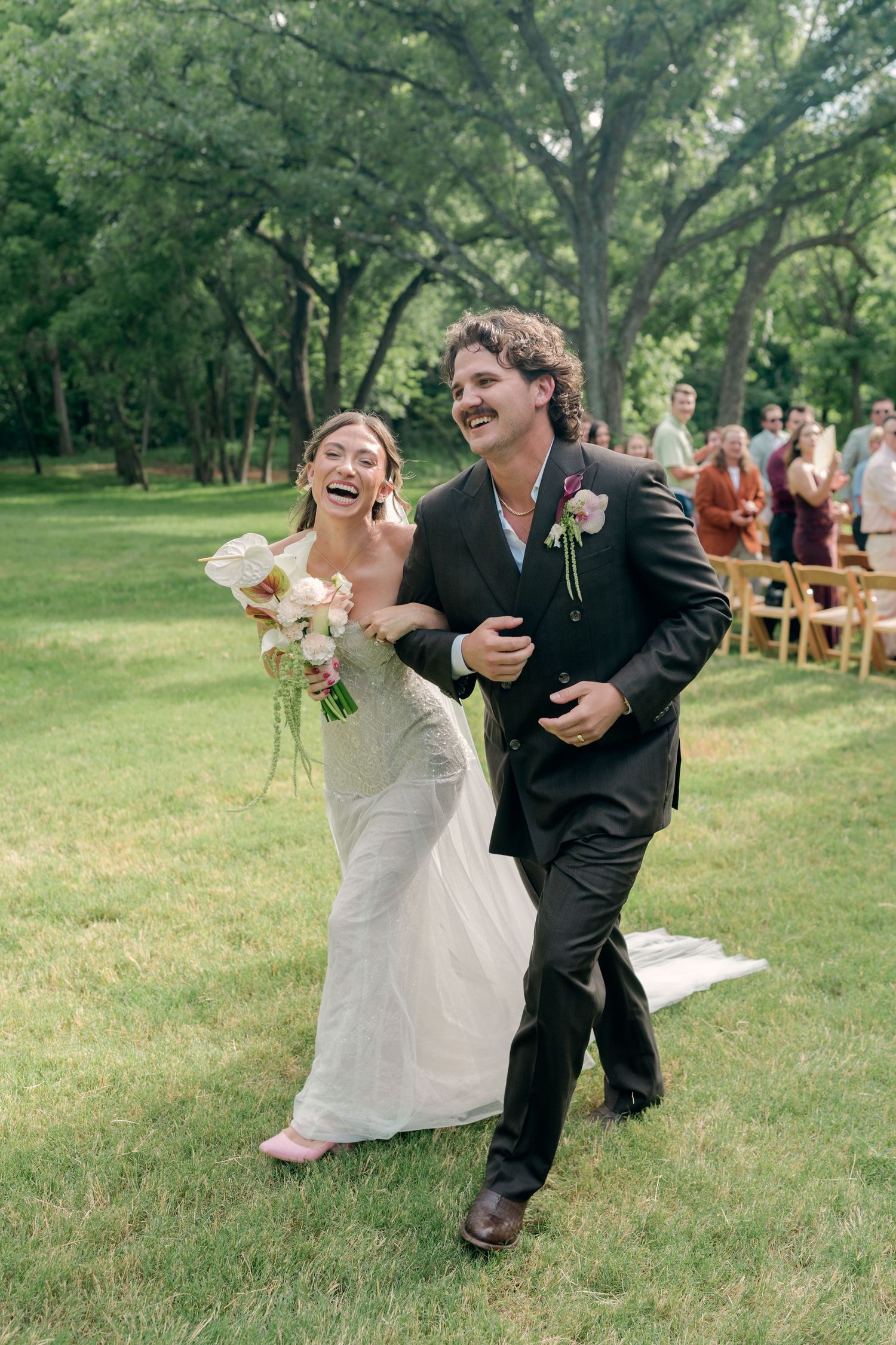 Joyful newlyweds walk down outdoor aisle laughing, bride in white gown with pink heels holding bouquet.