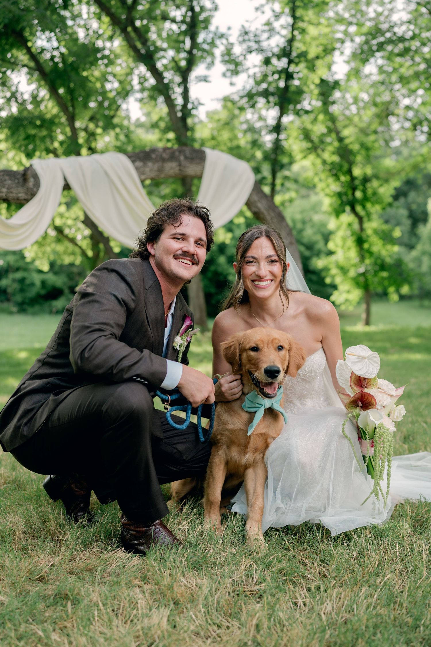 Smiling bride and groom crouch beside their golden retriever wearing a blue bow tie at outdoor wedding.
