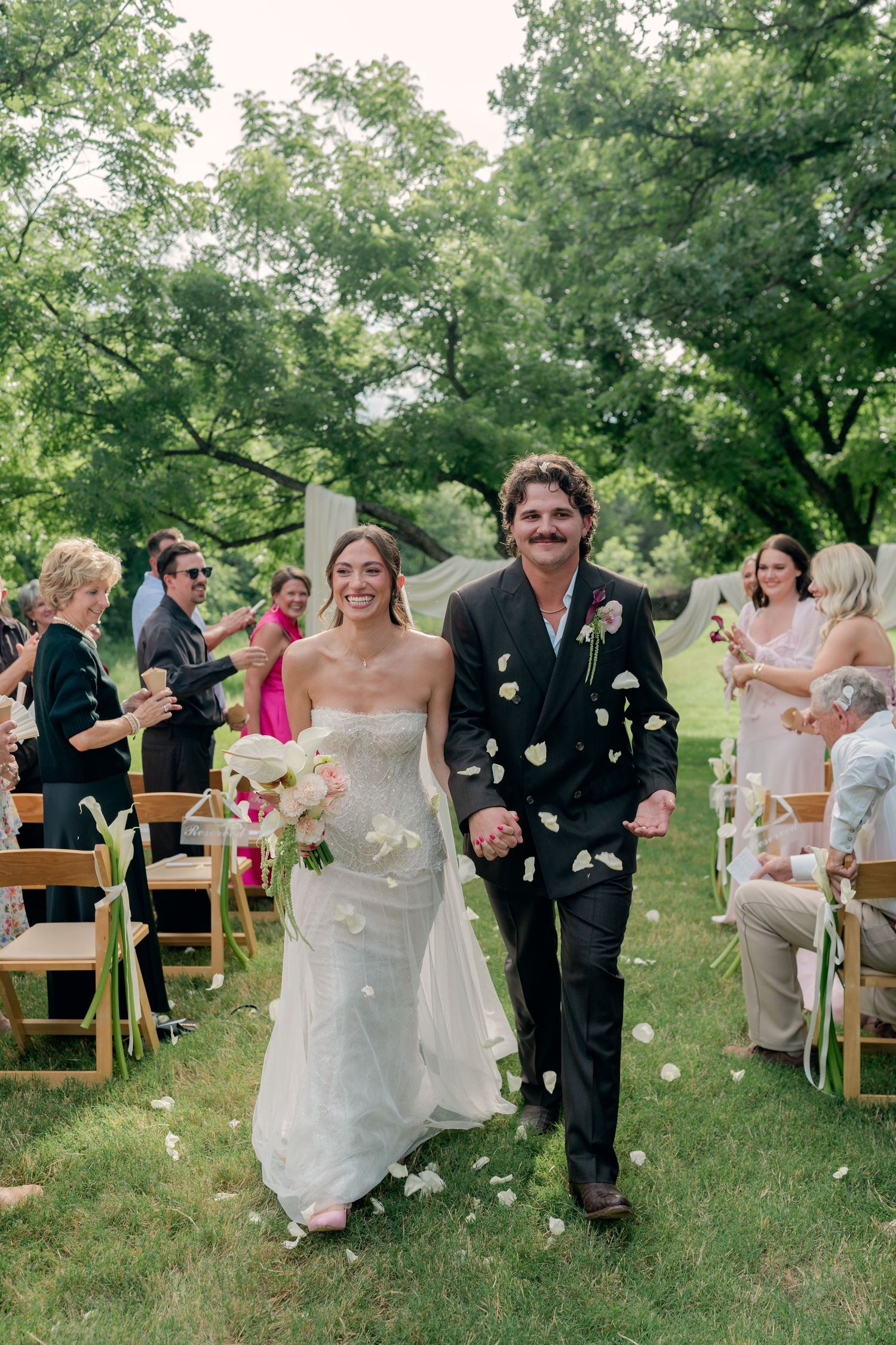 Bride and groom walk down outdoor wedding aisle as guests toss white flower petals in lush green garden setting.