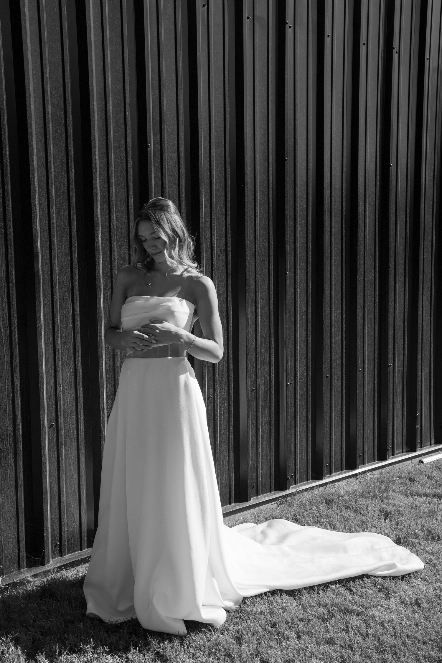 Bride in elegant white wedding gown poses against dark metal wall in black and white photo.