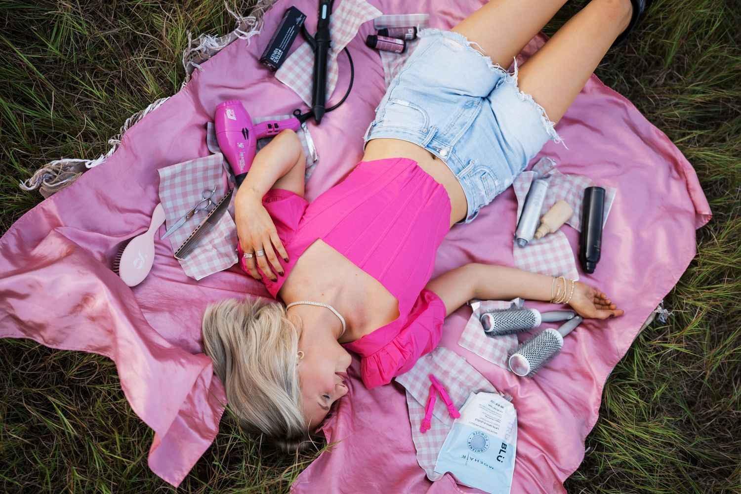 Someone lying on a pink blanket in the grass wearing denim shorts and a bright pink top with scattered items nearby.