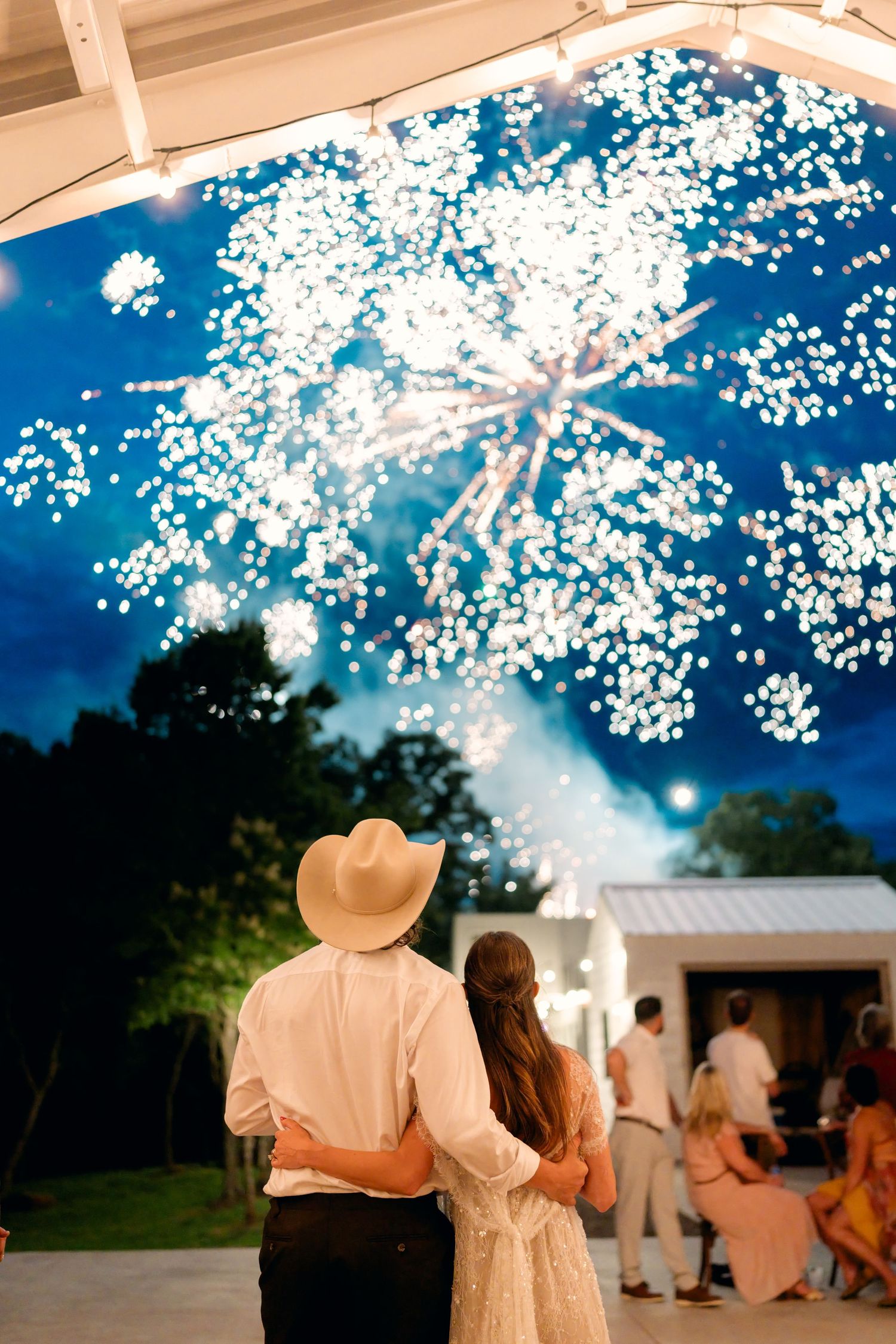 Bride and groom in cowboy hat watching fireworks display light up the night sky at wedding reception.