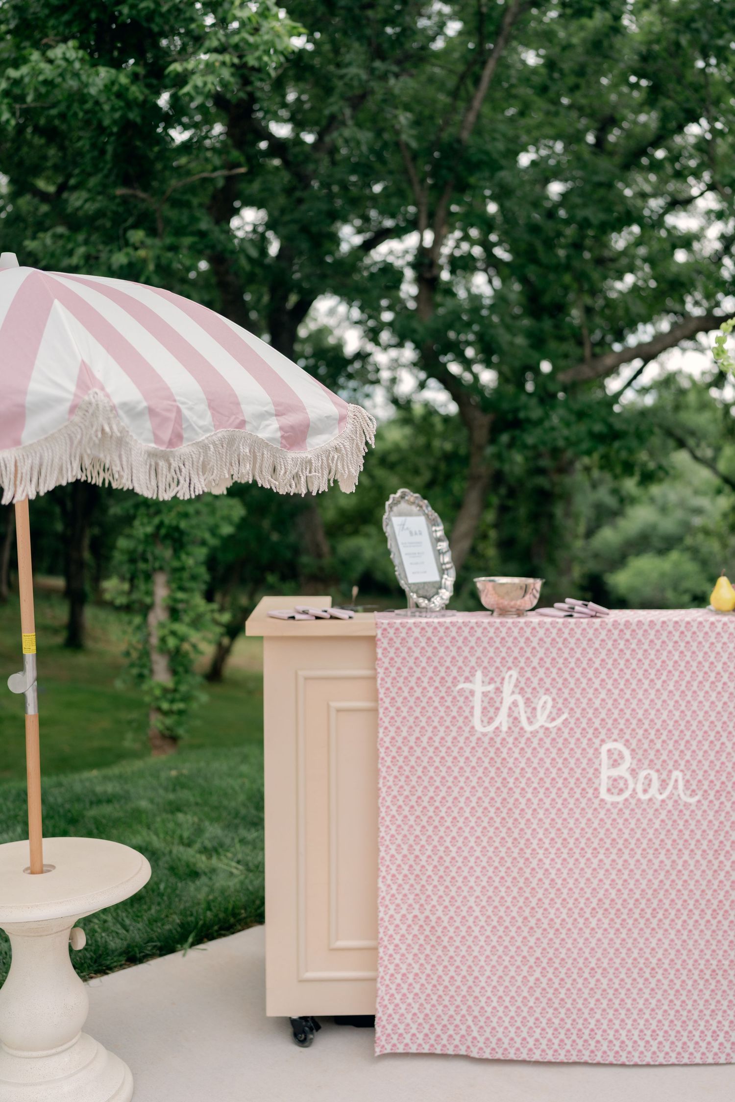 Pink outdoor wedding bar with striped fringed umbrella and elegant signage in a lush garden setting.