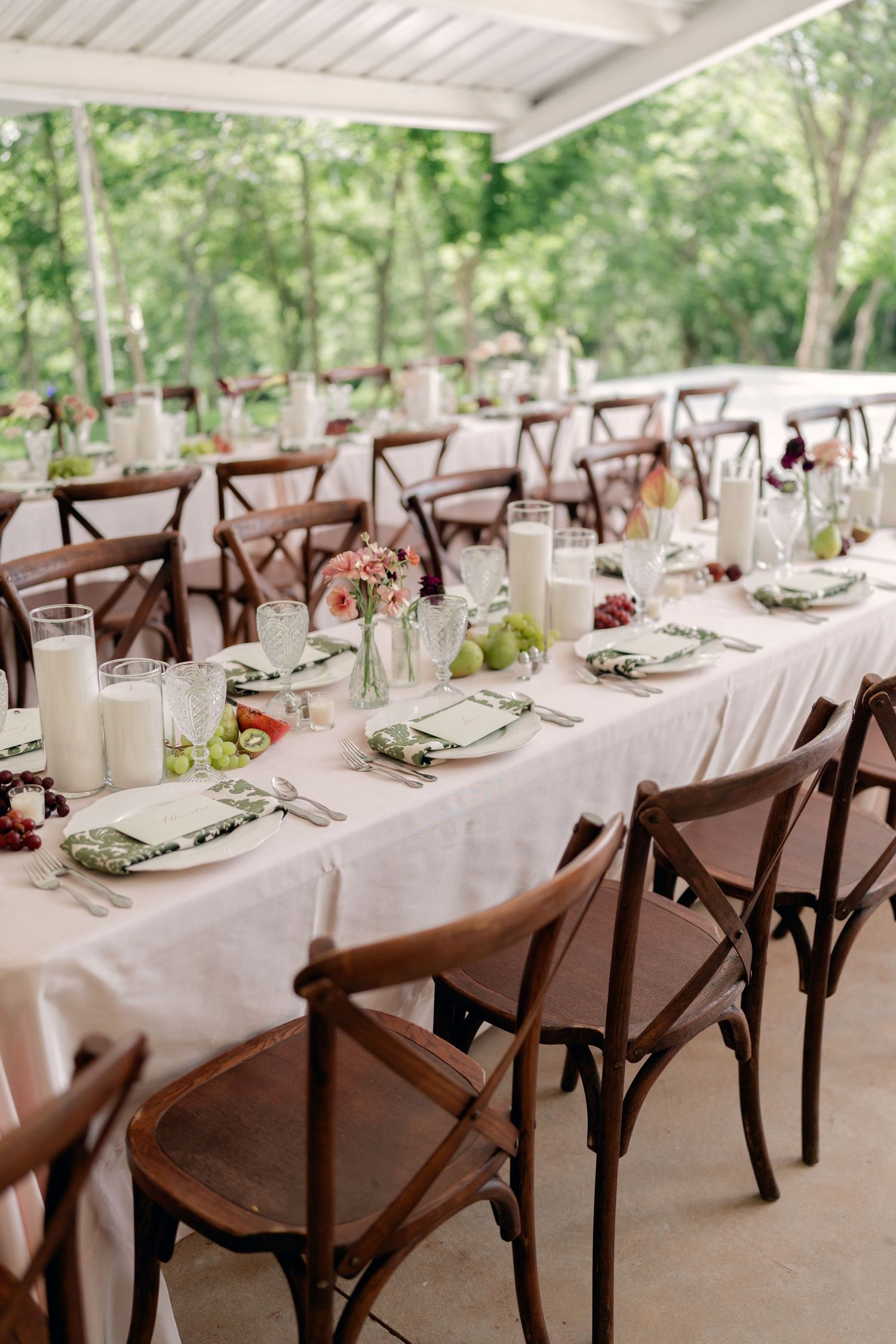 Elegant outdoor wedding reception with long tables, wooden cross-back chairs, candles, and floral centerpieces.