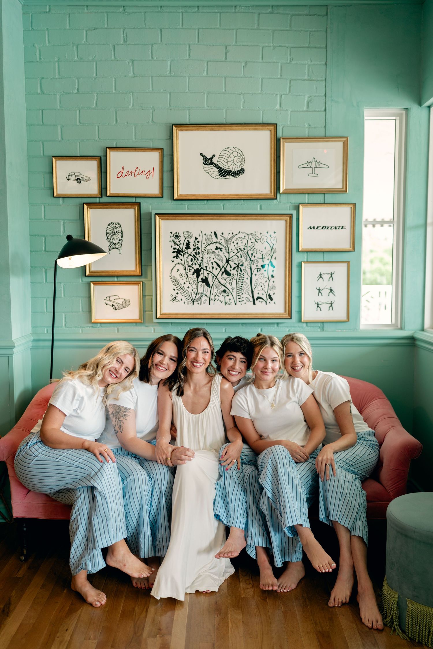 Bride and bridesmaids in white tops and blue striped pants sitting on a pink sofa with gallery wall backdrop.